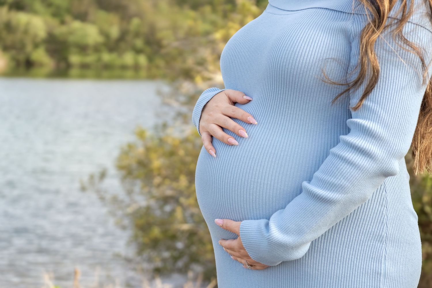 Pregnant woman in blue ribbed dress cradling baby bump outdoors near a serene lake surrounded by trees.