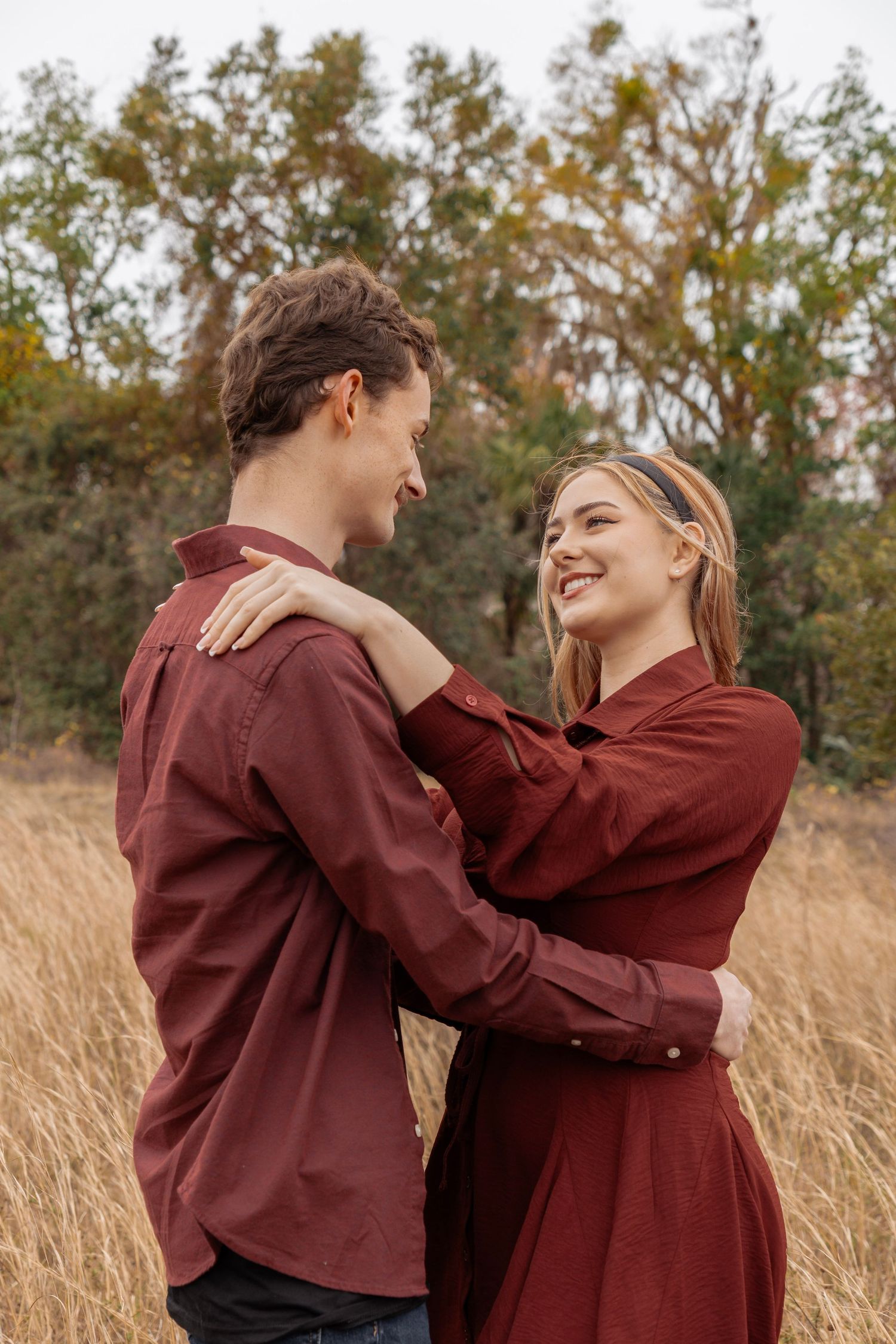 A smiling couple in matching burgundy outfits embrace in a golden field with autumn trees in the background.