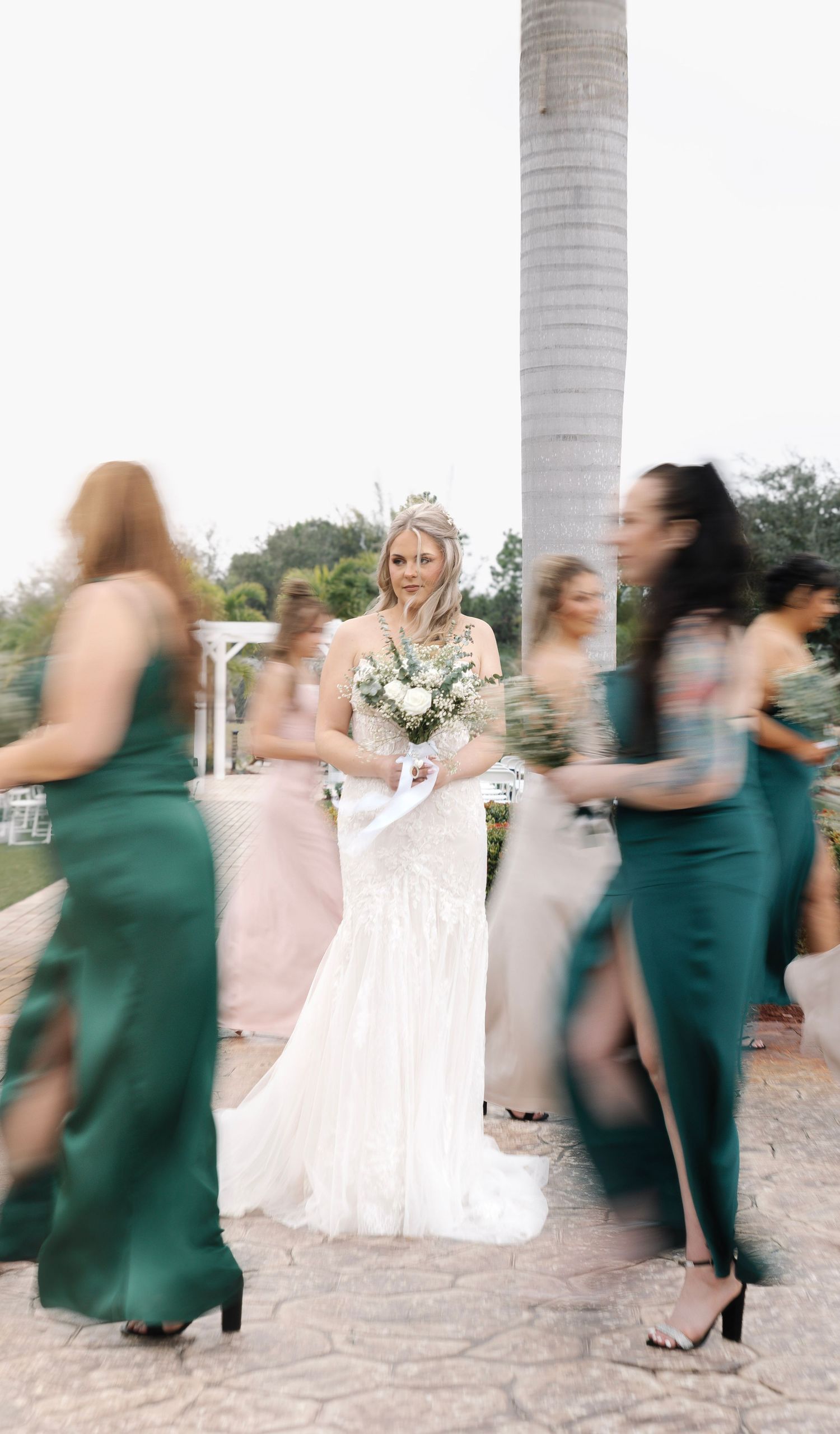 Bride in lace gown holds bouquet as bridesmaids in emerald dresses move around her outdoors.
