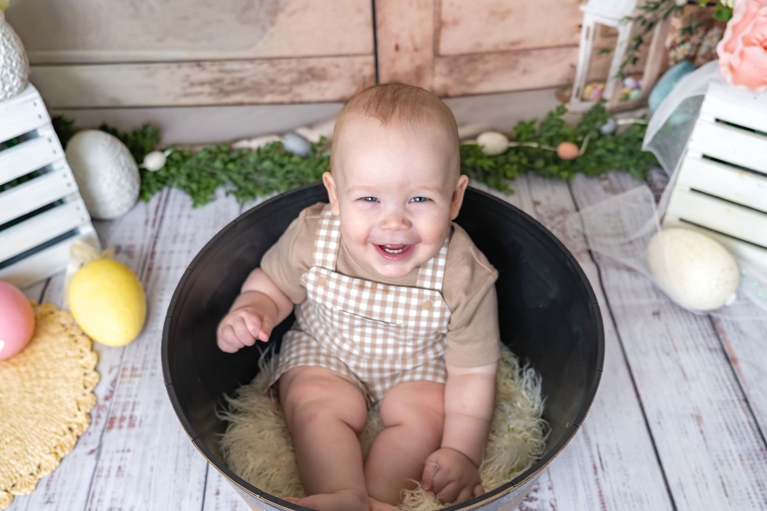 Smiling baby in a metal tub with Easter eggs and greenery decor, wearing a checkered romper for a festive photo.