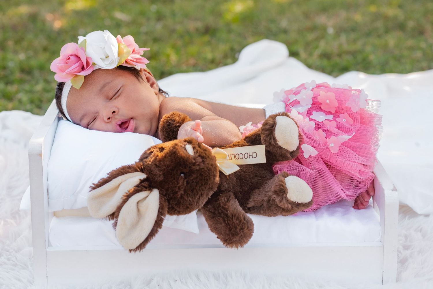 Sleeping newborn baby girl in pink tutu skirt and floral headband cuddling a brown teddy bear in a white wooden crate.