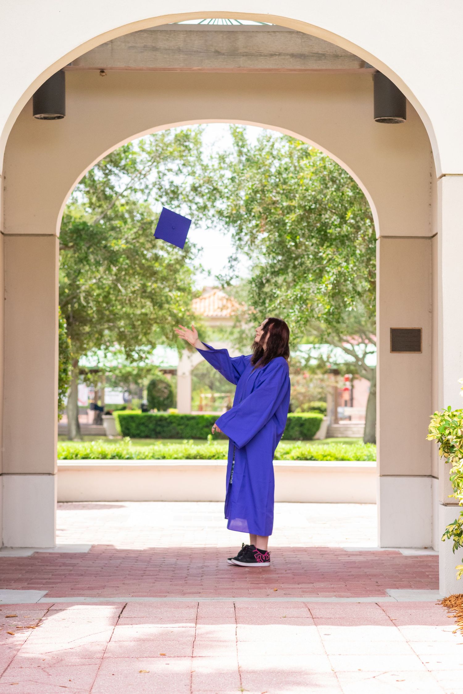 Graduate in blue cap and gown tossing mortarboard under archway on campus.