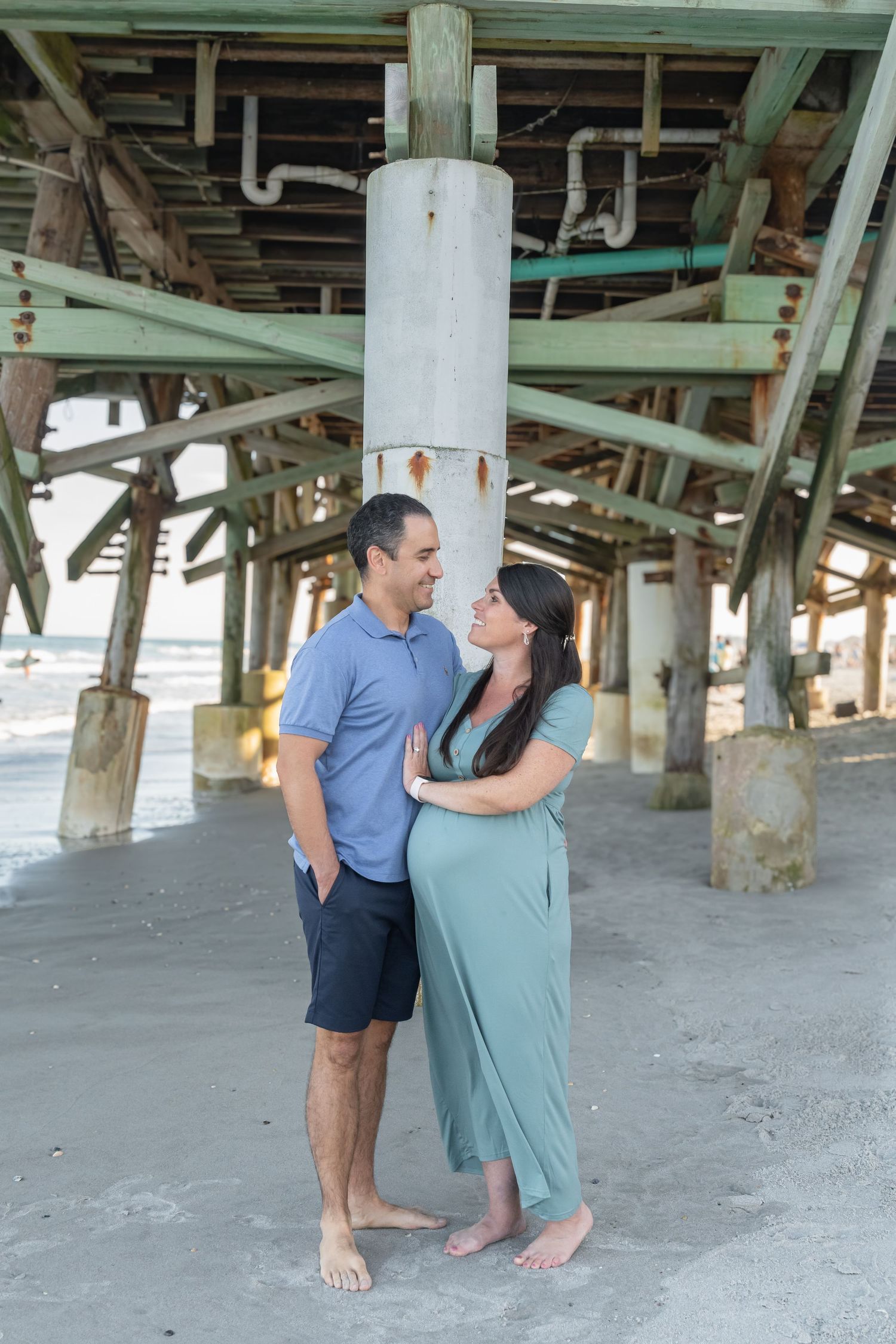Pregnant couple embracing under a beach pier, woman in mint dress, man in blue shirt, barefoot on sand.