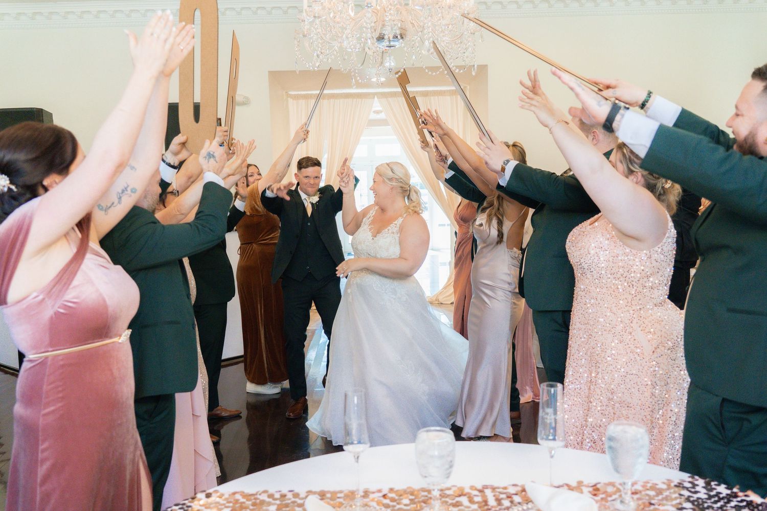 Wedding party raises sparklers and signs to celebrate bride and groom's joyful reception entrance indoors.
