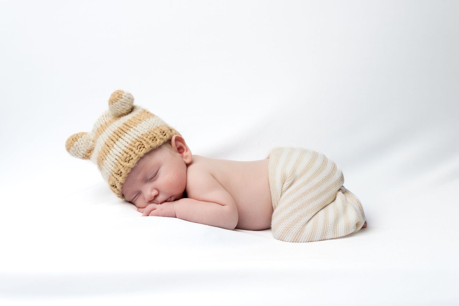 Sleeping newborn baby wearing a knitted bear ear hat and striped pants on a white background.