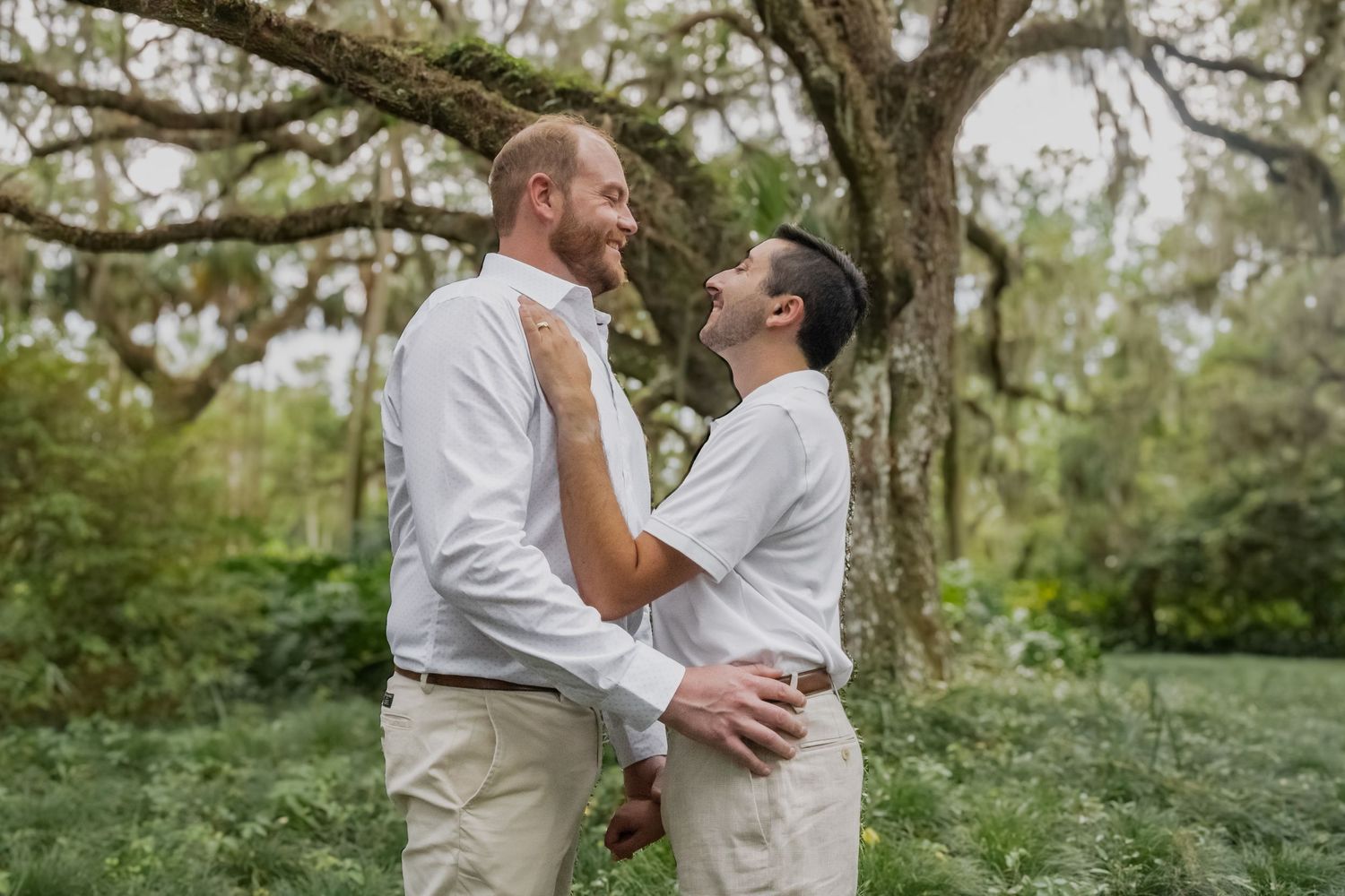 Two grooms in white shirts embrace and smile at each other outdoors under a moss-covered oak tree.