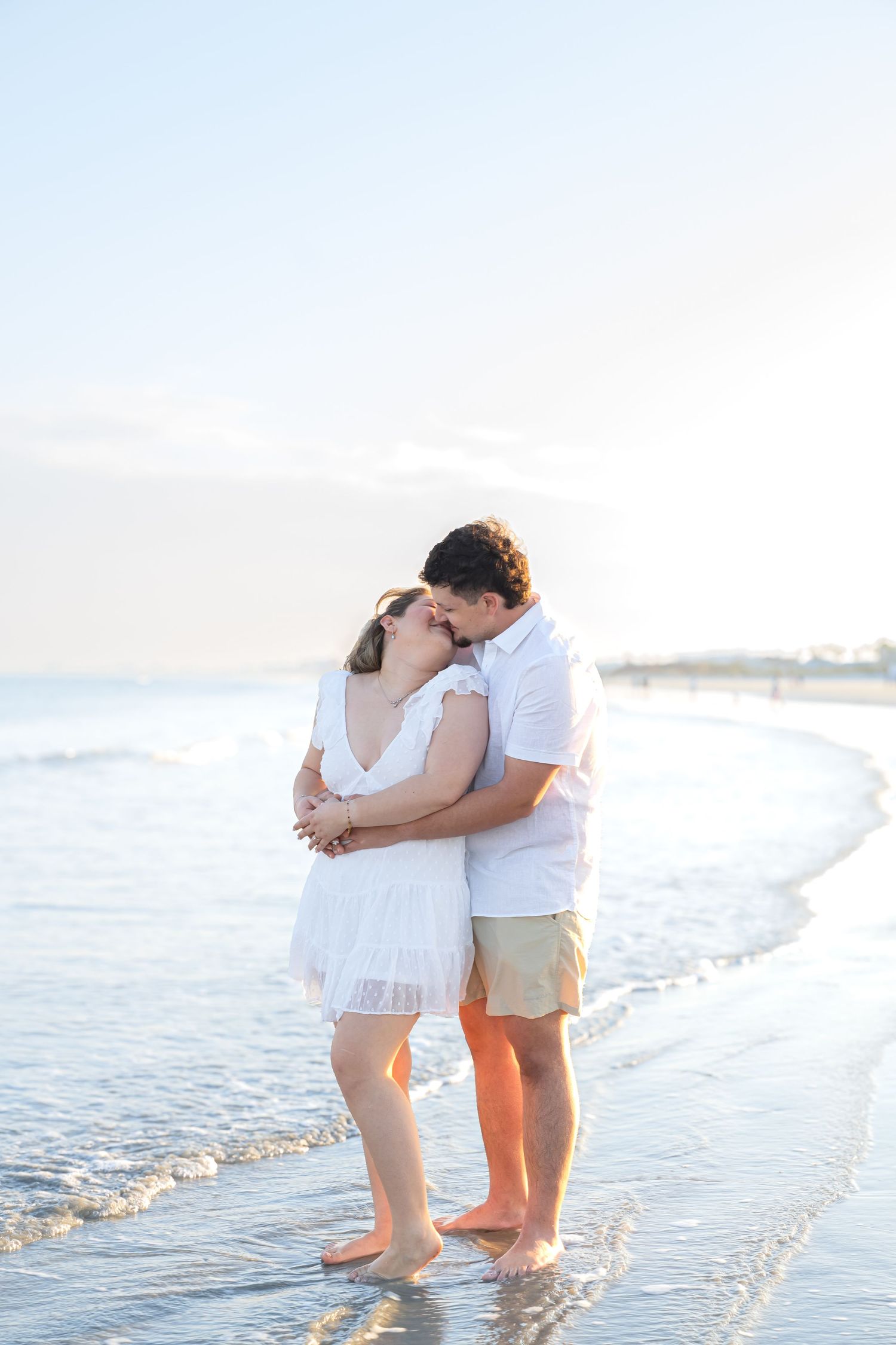 Couple embracing and kissing on a sunny beach, wearing white outfits, standing in shallow ocean waves.