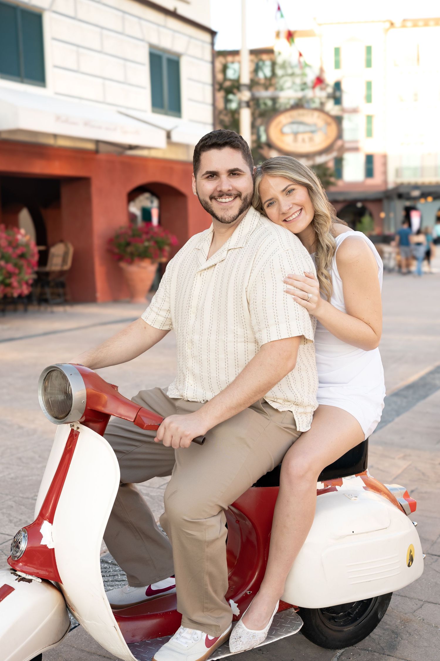 Smiling couple posing together on a classic red and white Vespa scooter in a town square.