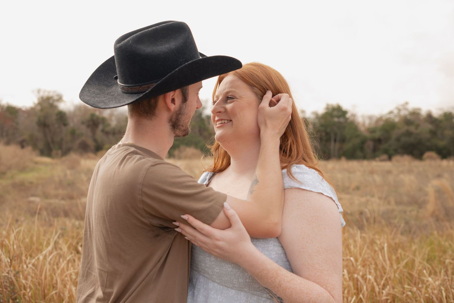 Couple in a golden field, man in black cowboy hat and brown shirt, woman in white dress, sharing a romantic moment.