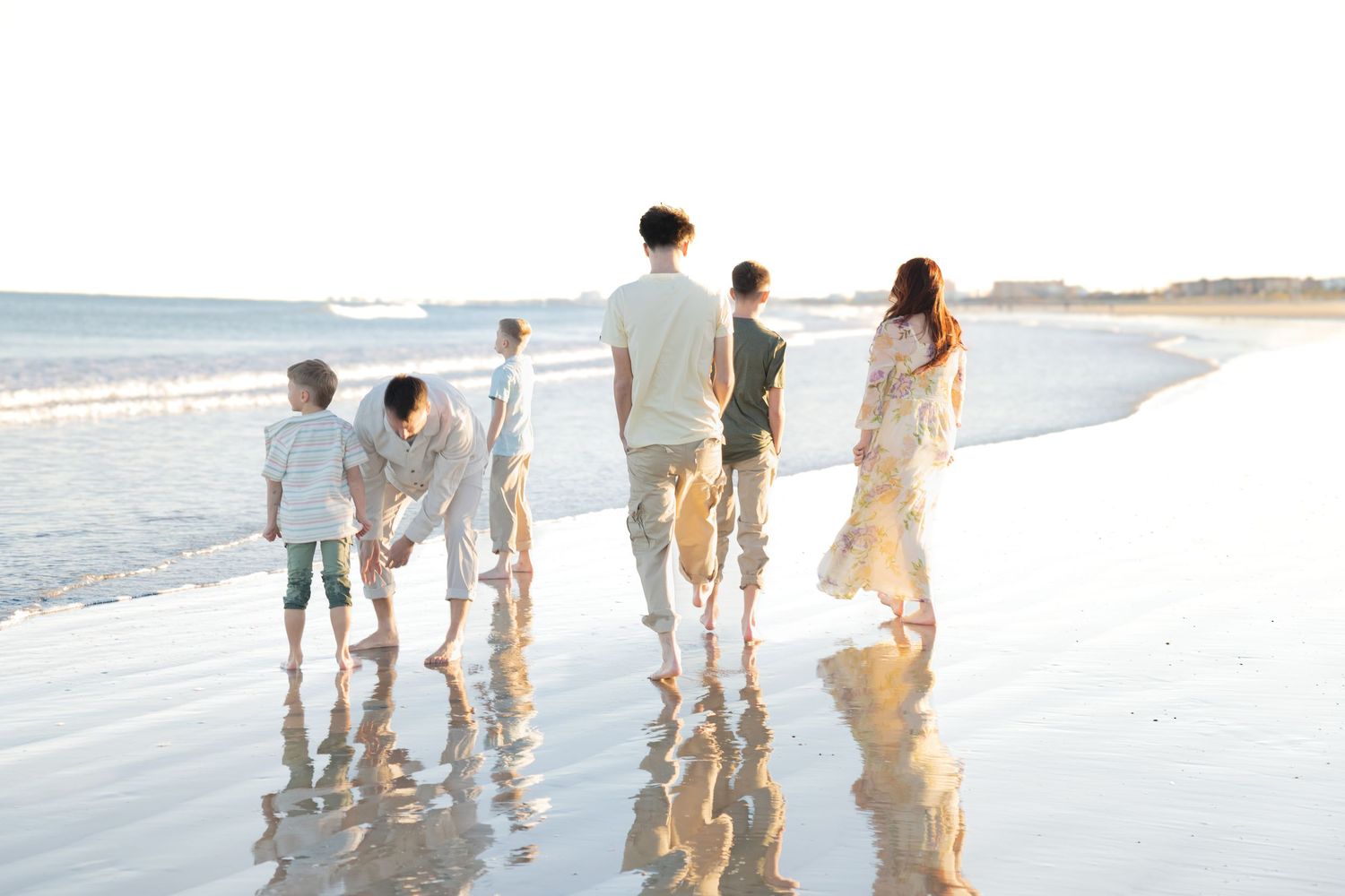 Family of six walking hand-in-hand along a sunlit beach shoreline with reflections on wet sand.