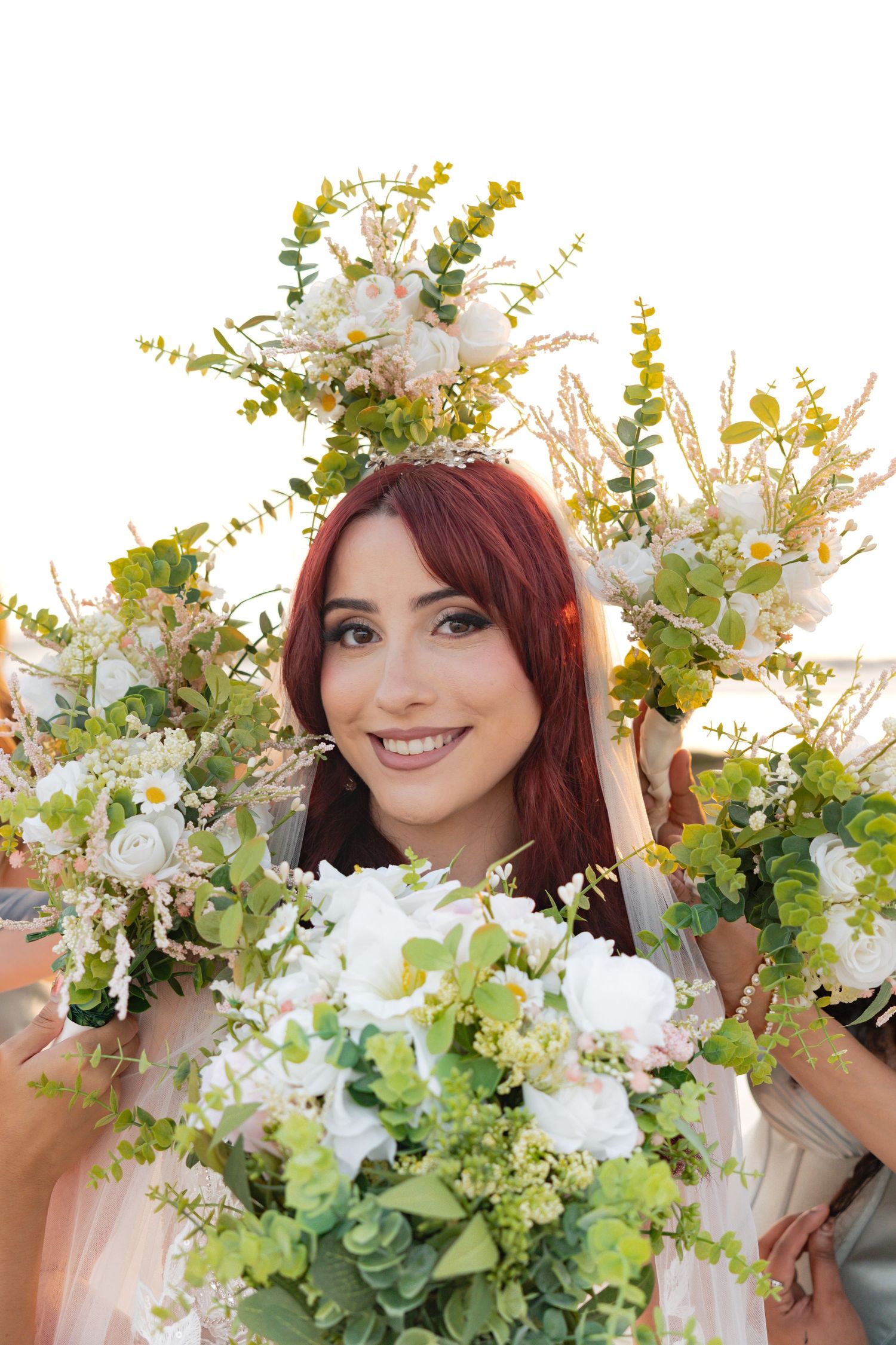 Smiling bride with red hair wearing a floral crown, surrounded by white and green wedding bouquets.