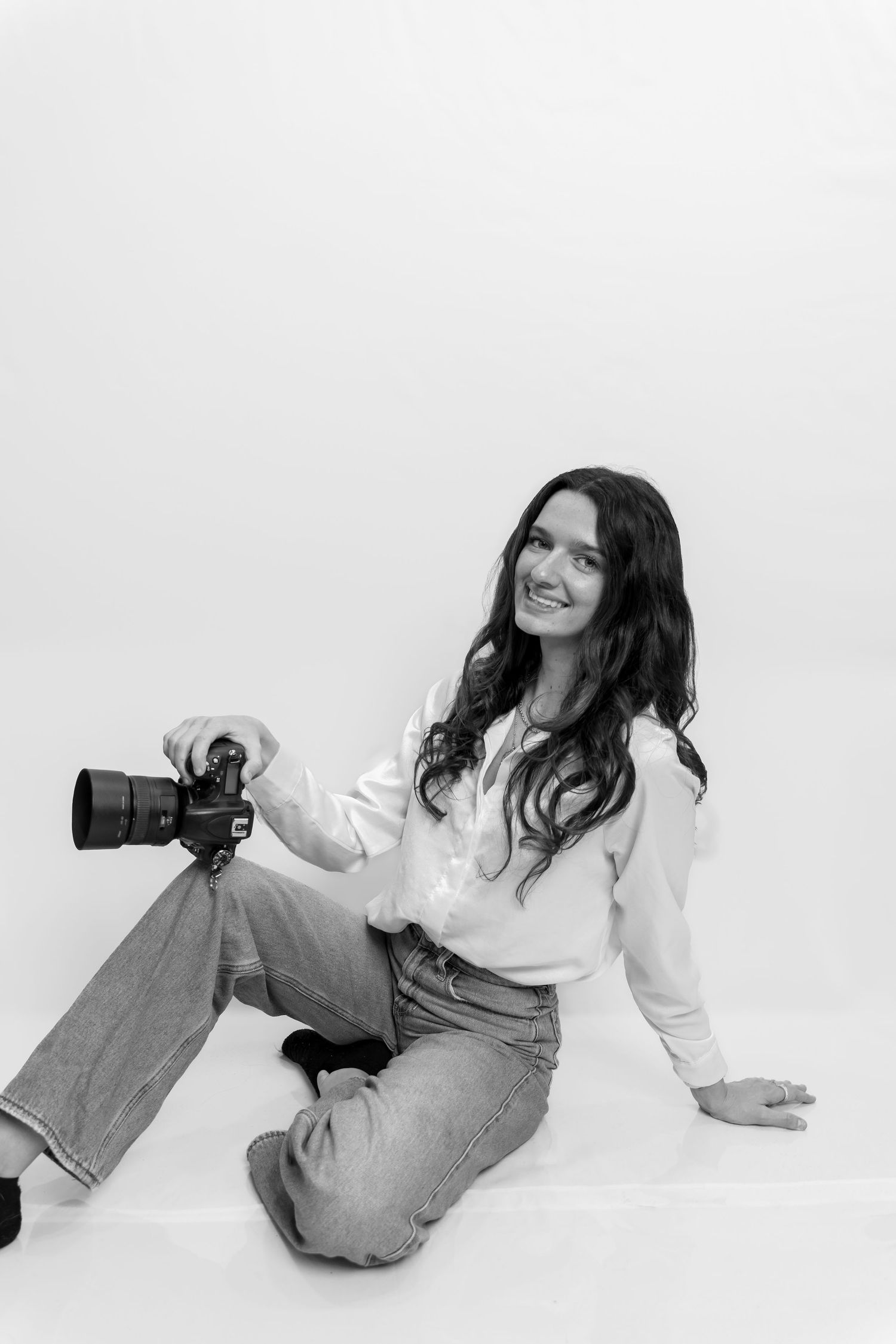Black and white photo of a smiling woman sitting on floor holding a DSLR camera, wearing white shirt and jeans.