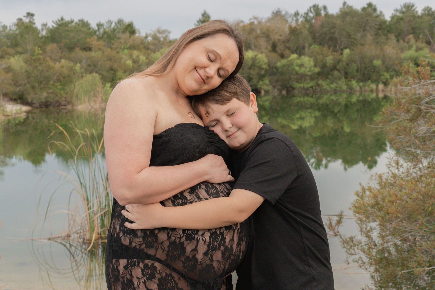 Pregnant woman in black lace dress embraced by child near a serene lake with lush greenery backdrop.