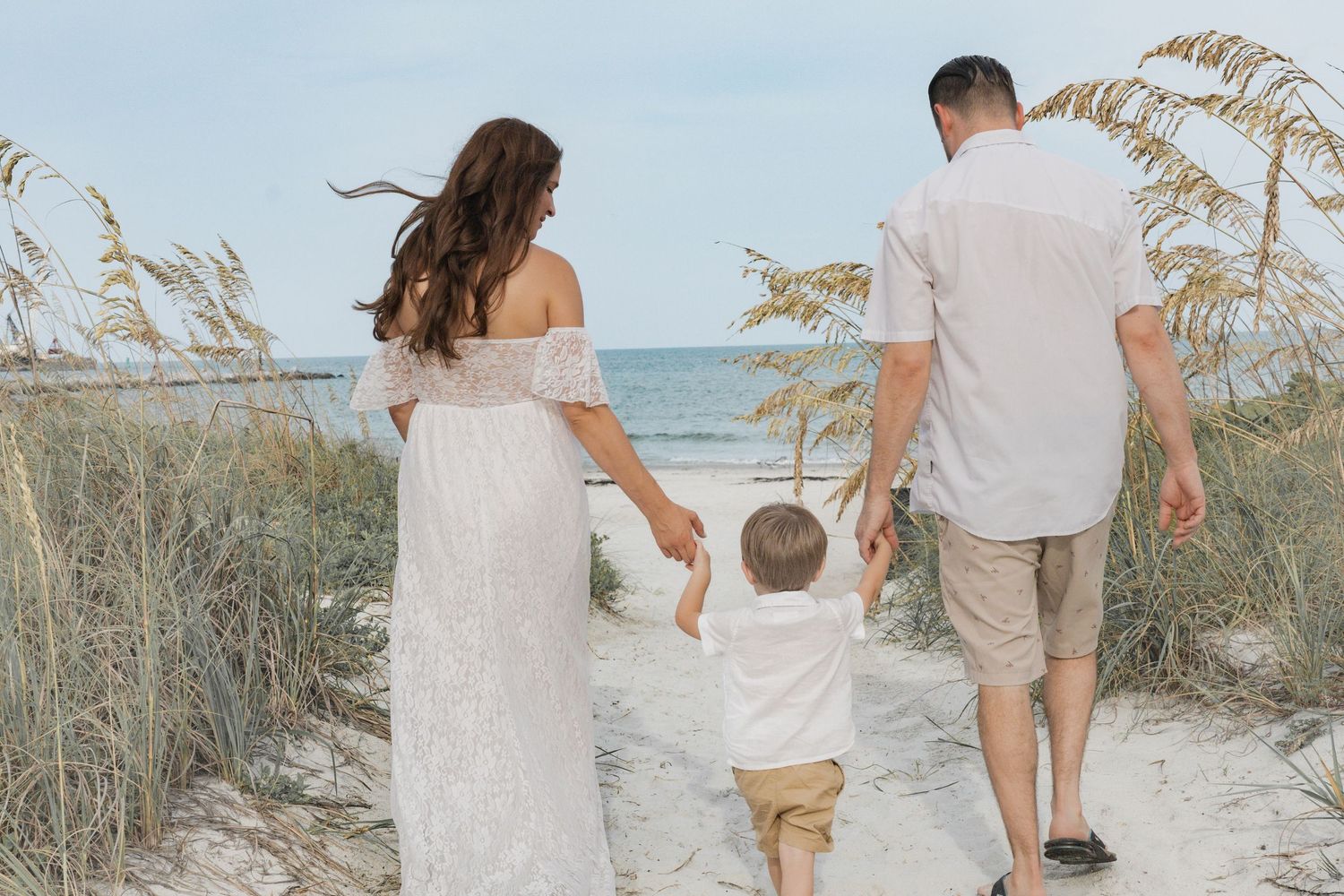 Family maternity photo with pregnant woman in white lace dress walking to beach with husband and son.