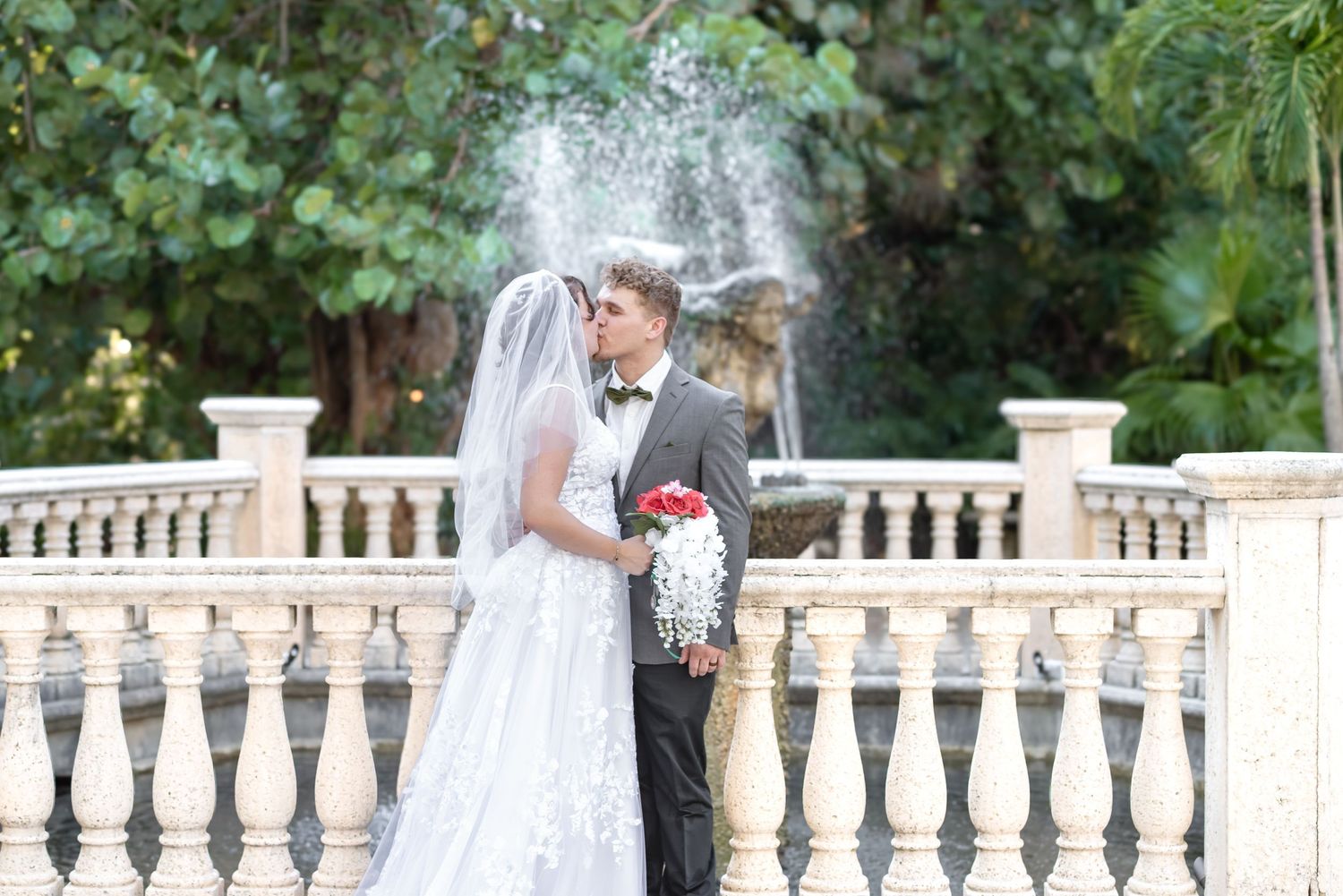 Bride and groom share a romantic moment by a stone balustrade with a fountain and lush greenery backdrop.