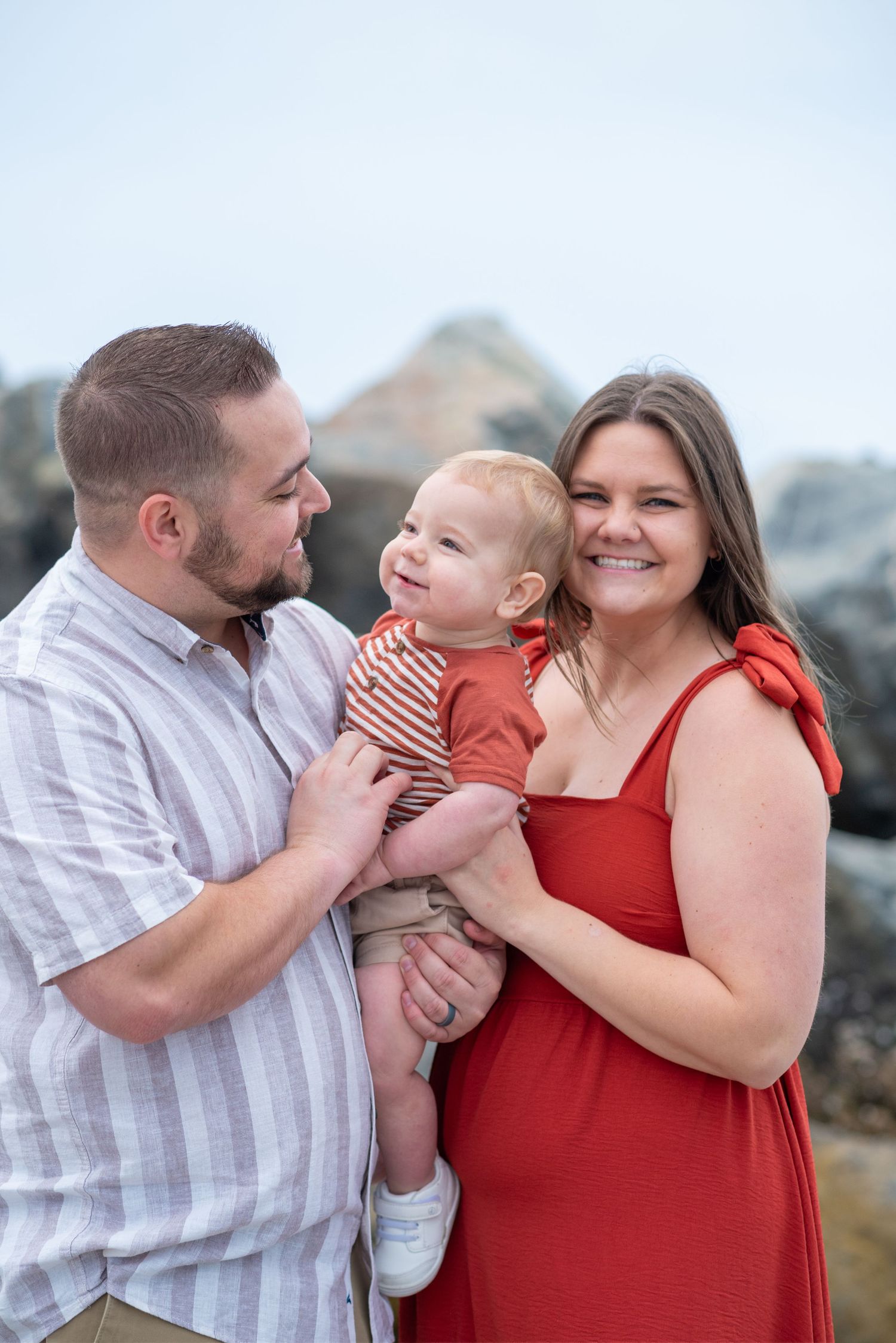 A happy family of three poses outdoors, with parents holding their smiling baby near rocky shores.