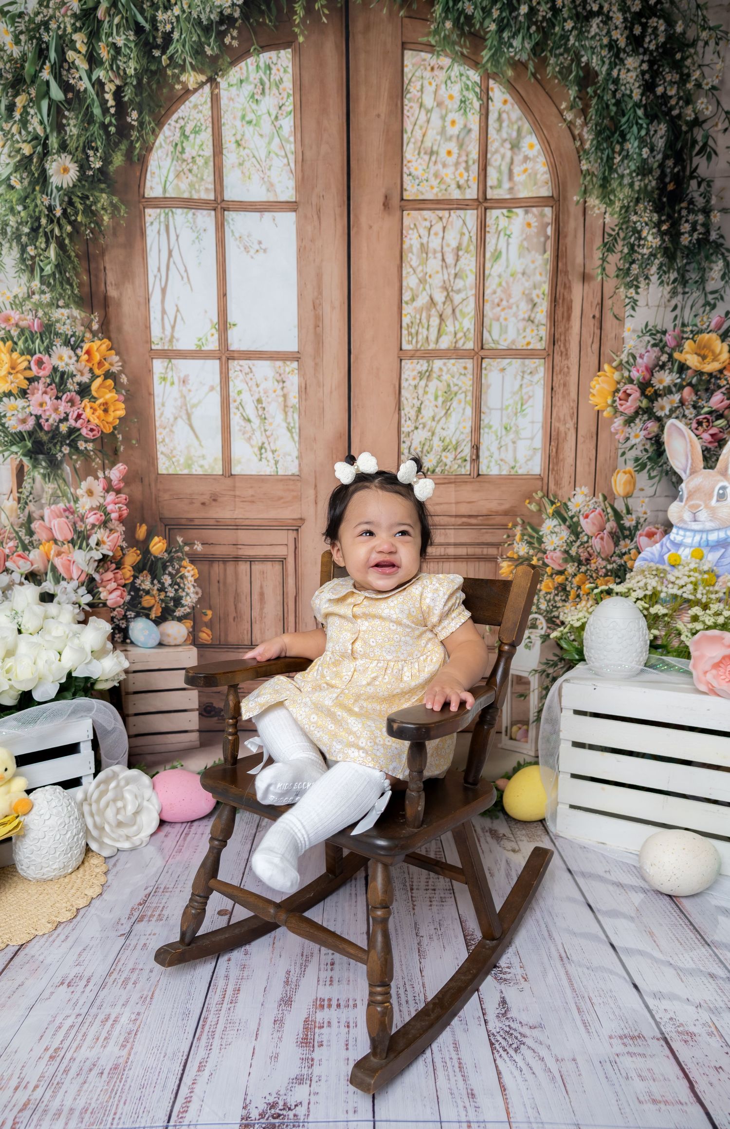 Smiling baby girl in yellow dress sits in wooden rocking chair amid Easter floral backdrop with bunny decor.