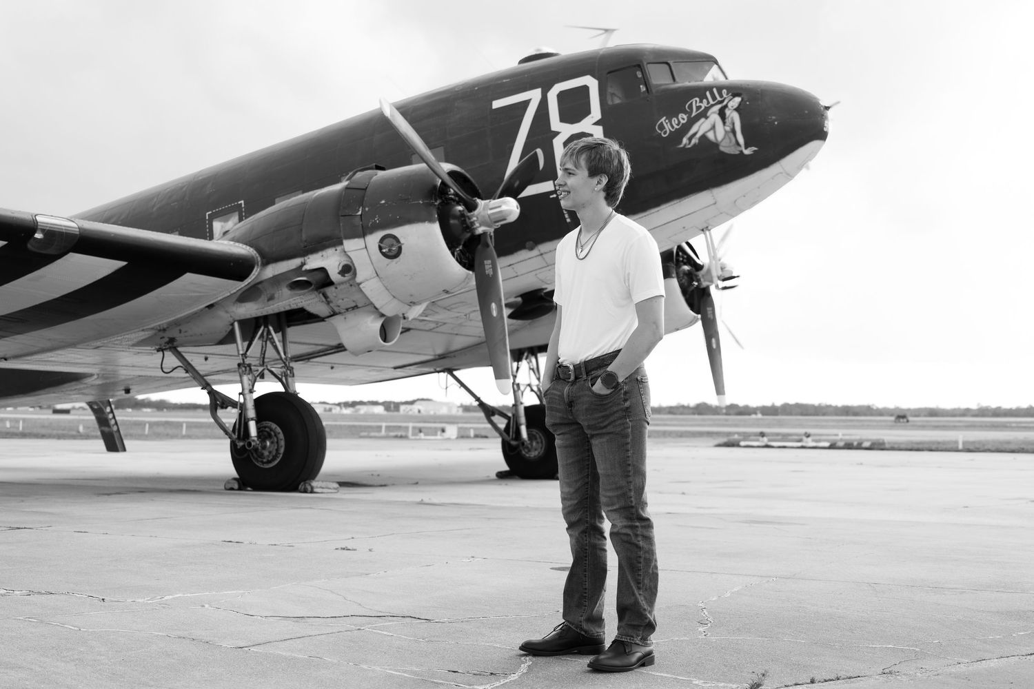 Black and white photo of teen boy posing beside vintage WWII propeller aircraft numbered 78.