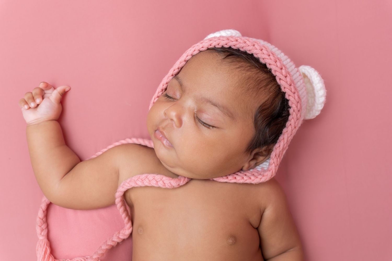 Sleeping newborn baby wearing a pink knit bear bonnet hat on a pink background.