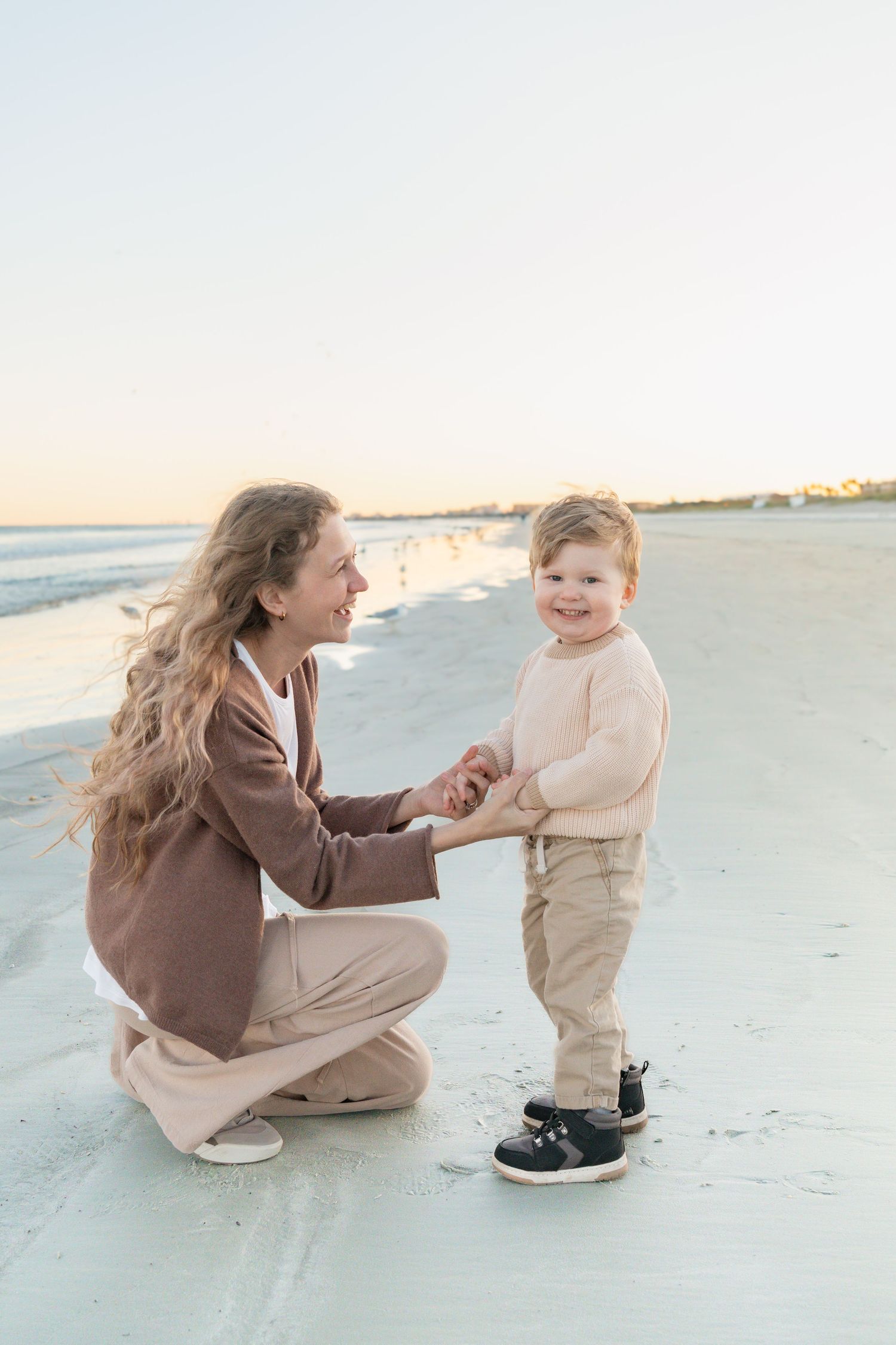 A smiling mother crouches on a sandy beach holding hands with her happy toddler son at sunset.