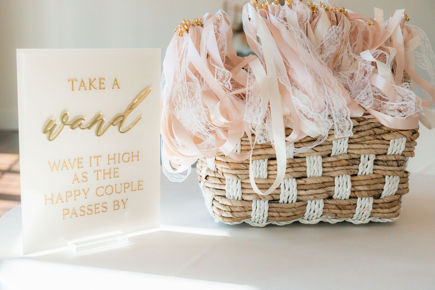 A wicker basket filled with blush pink ribbon wands beside a gold calligraphy sign for a wedding send-off.