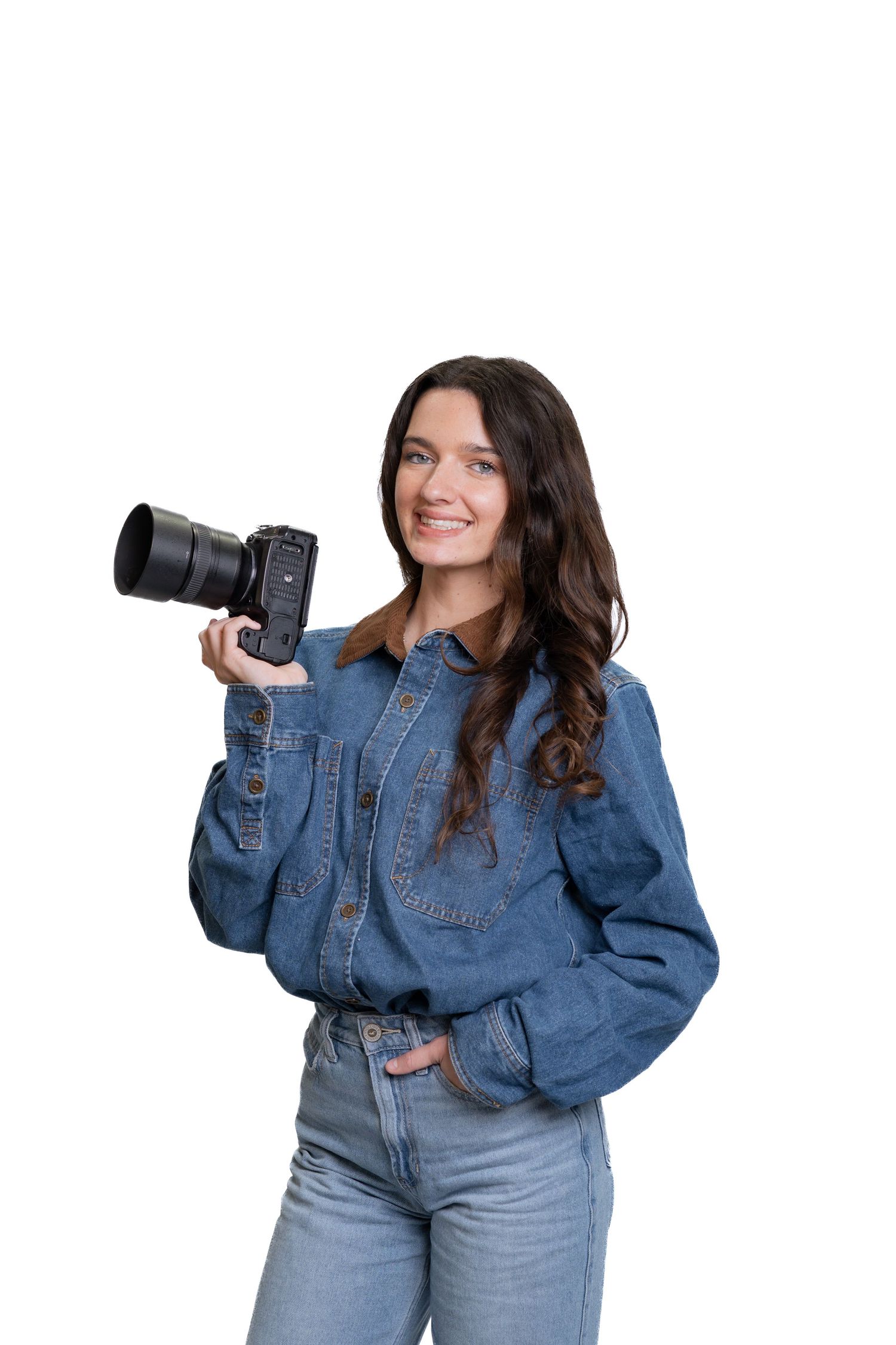 Young woman photographer in denim jacket holding professional DSLR camera, smiling against white background.