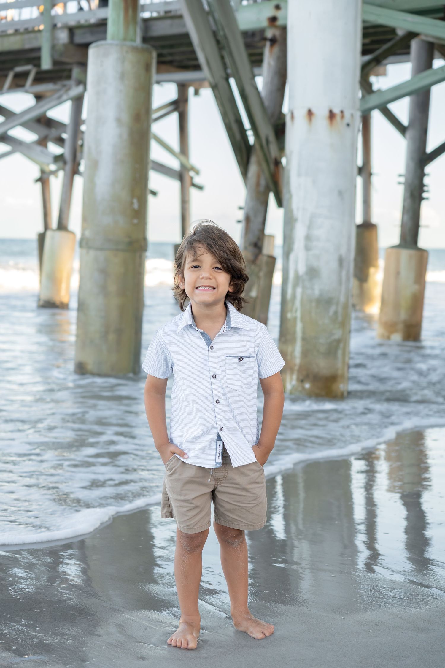 Smiling young boy in white shirt and khaki shorts standing barefoot under a beach pier.