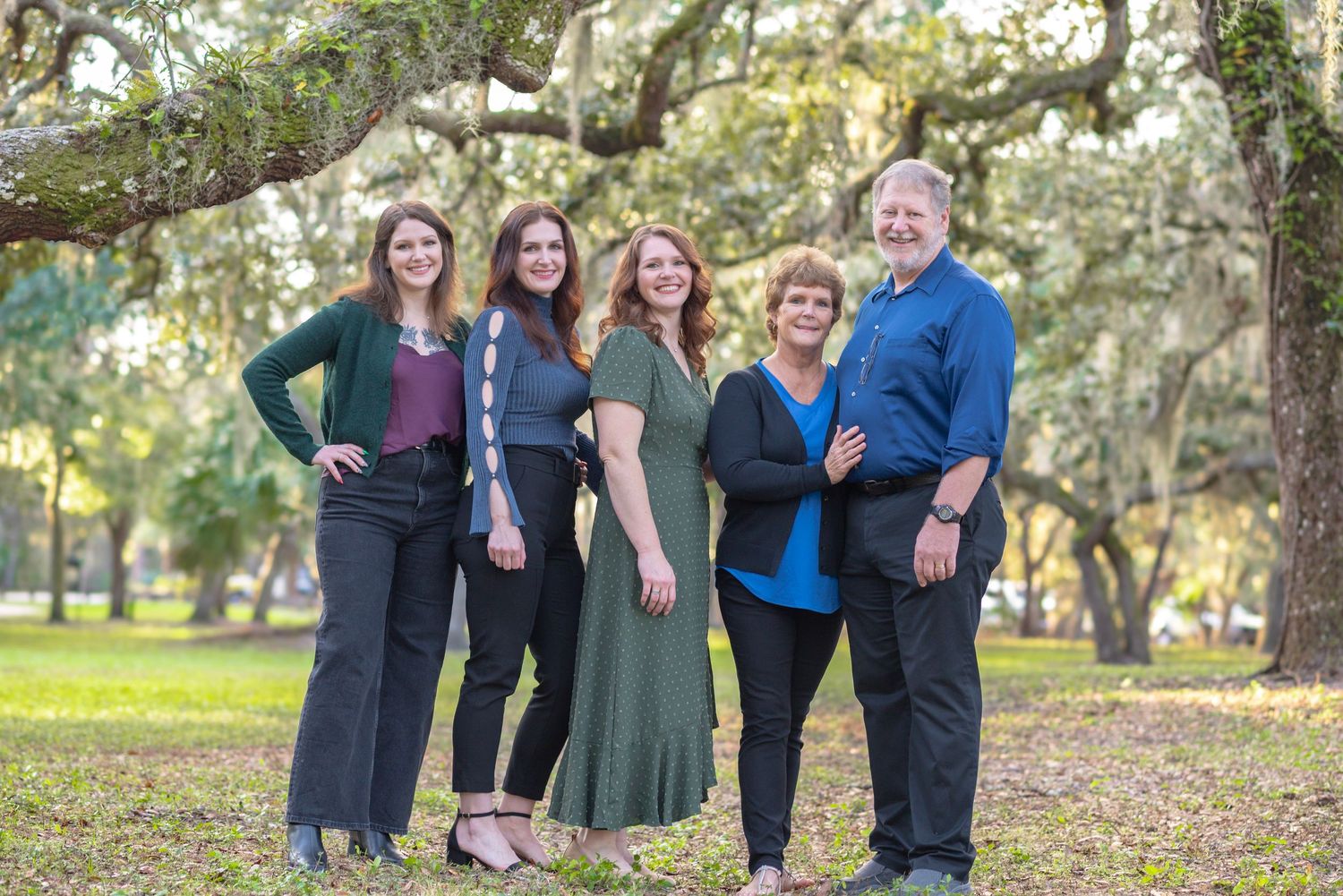 Family of five smiling together outdoors under oak trees in a park, dressed in elegant casual attire.