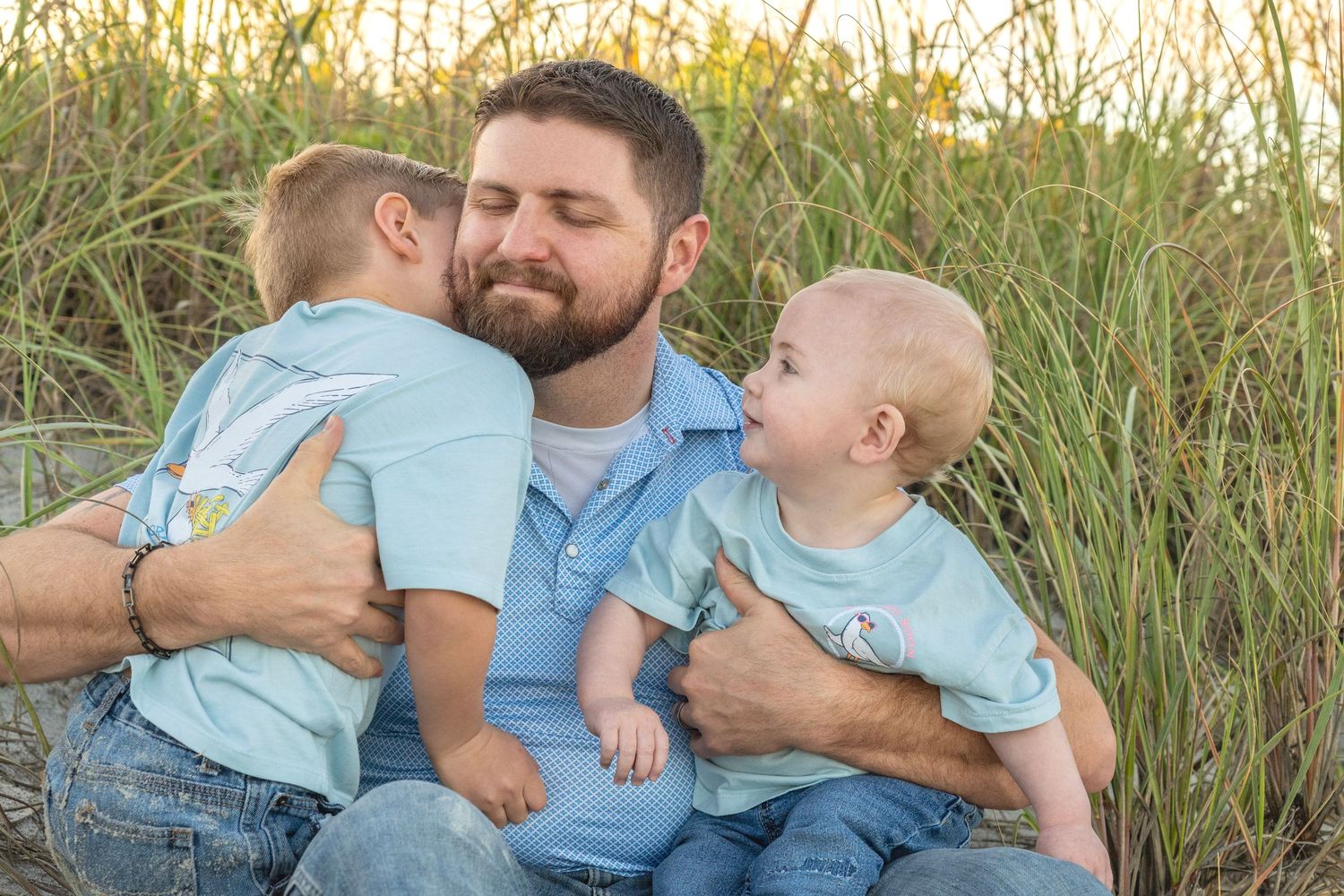 A bearded father sits in beach grass holding two young boys in matching light blue shirts, sharing a tender moment.