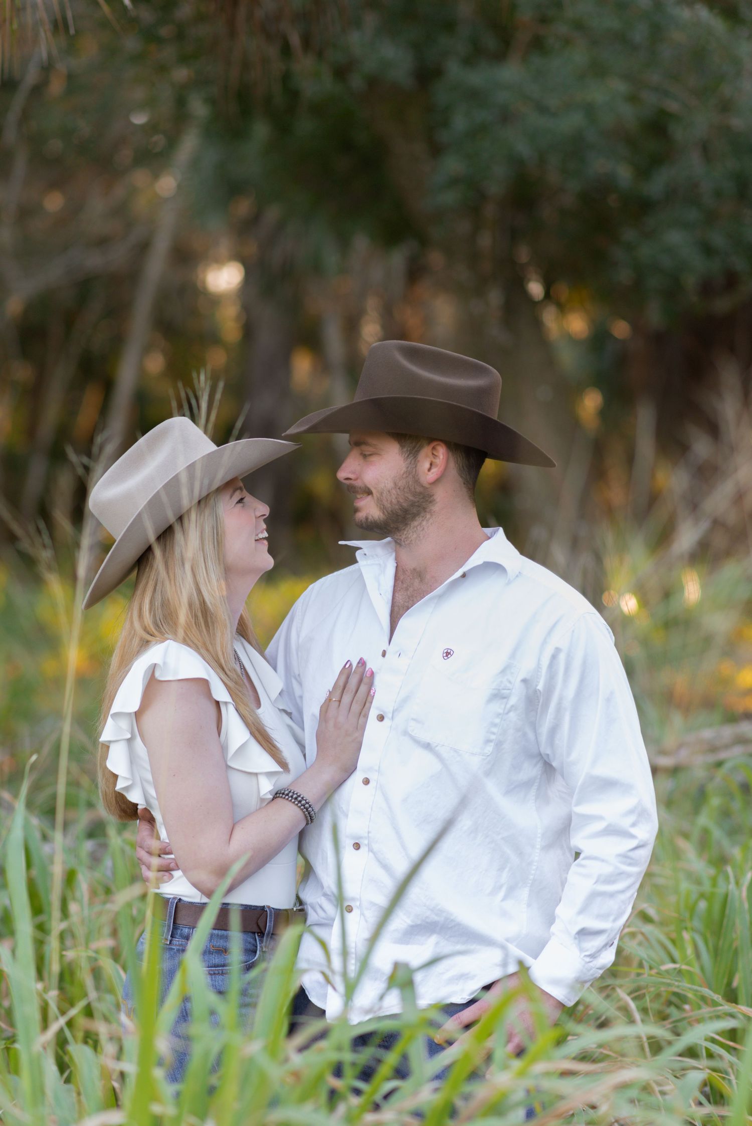 A couple in cowboy hats sharing a romantic moment outdoors, dressed in white tops and jeans among lush greenery.