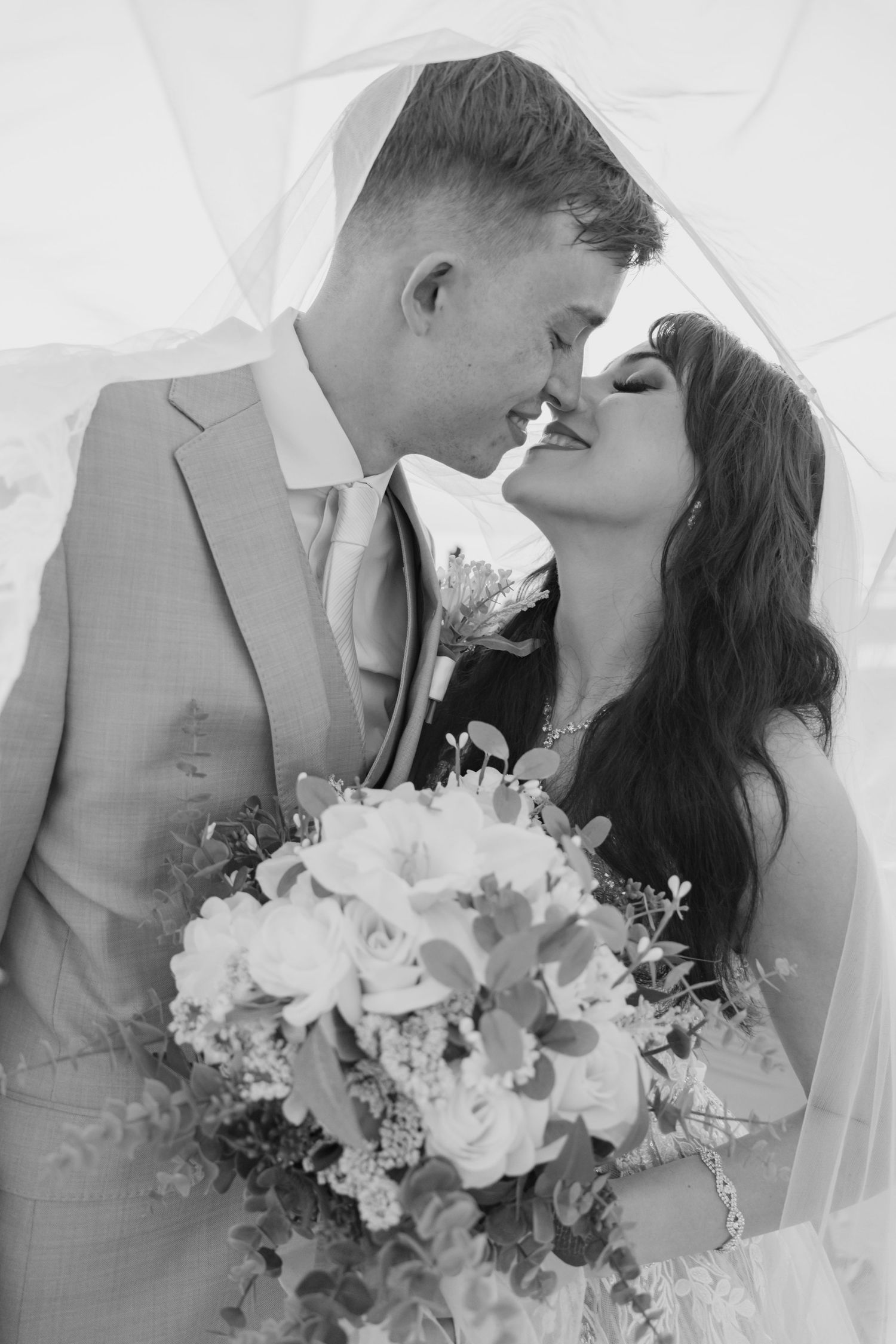 Black and white photo of bride and groom sharing romantic moment under veil holding white rose bouquet.