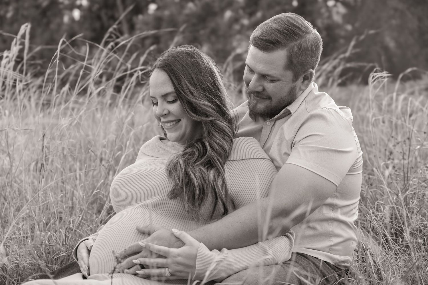 Black and white photo of a smiling couple sitting in a field, the man embracing the woman from behind.