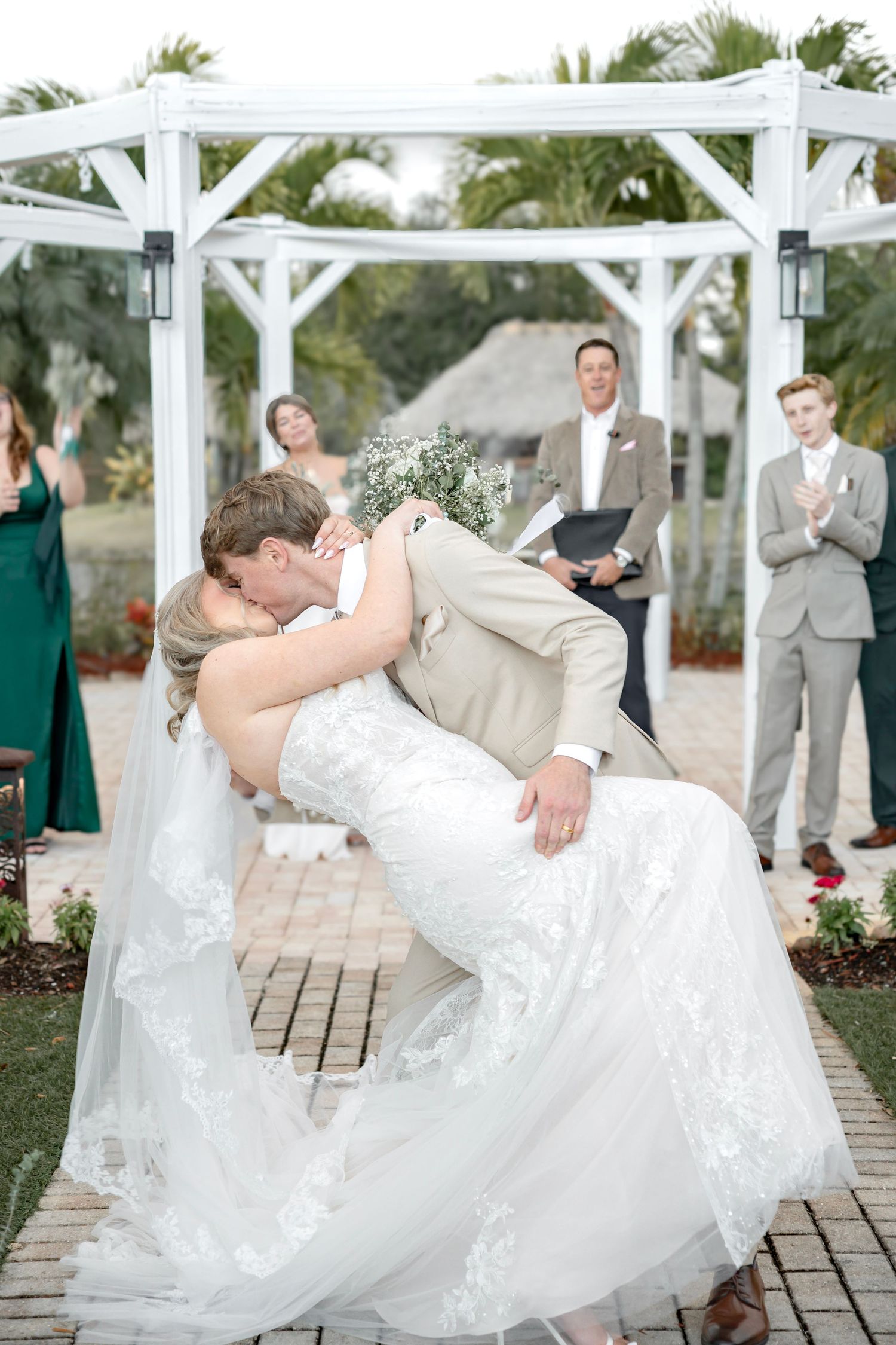 Bride and groom sharing a romantic dip kiss under a white pergola at outdoor ceremony.