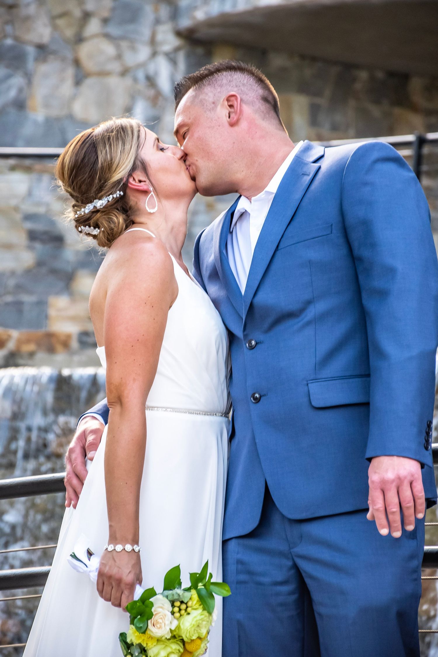 Bride and groom share a romantic kiss outdoors, bride holding yellow and white bouquet, groom in blue suit.
