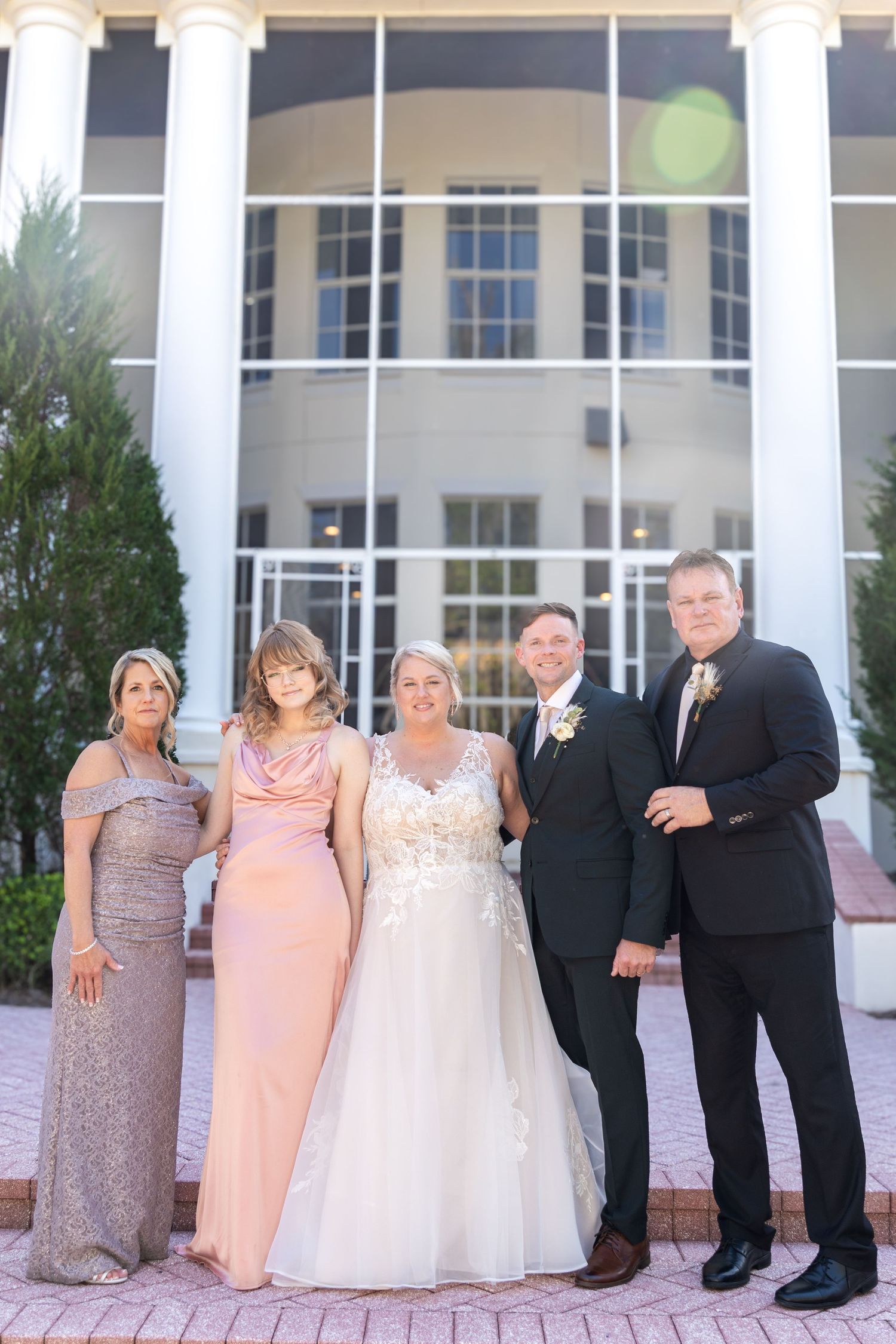 Bride and groom pose with family in formal attire outside a grand white columned wedding venue.