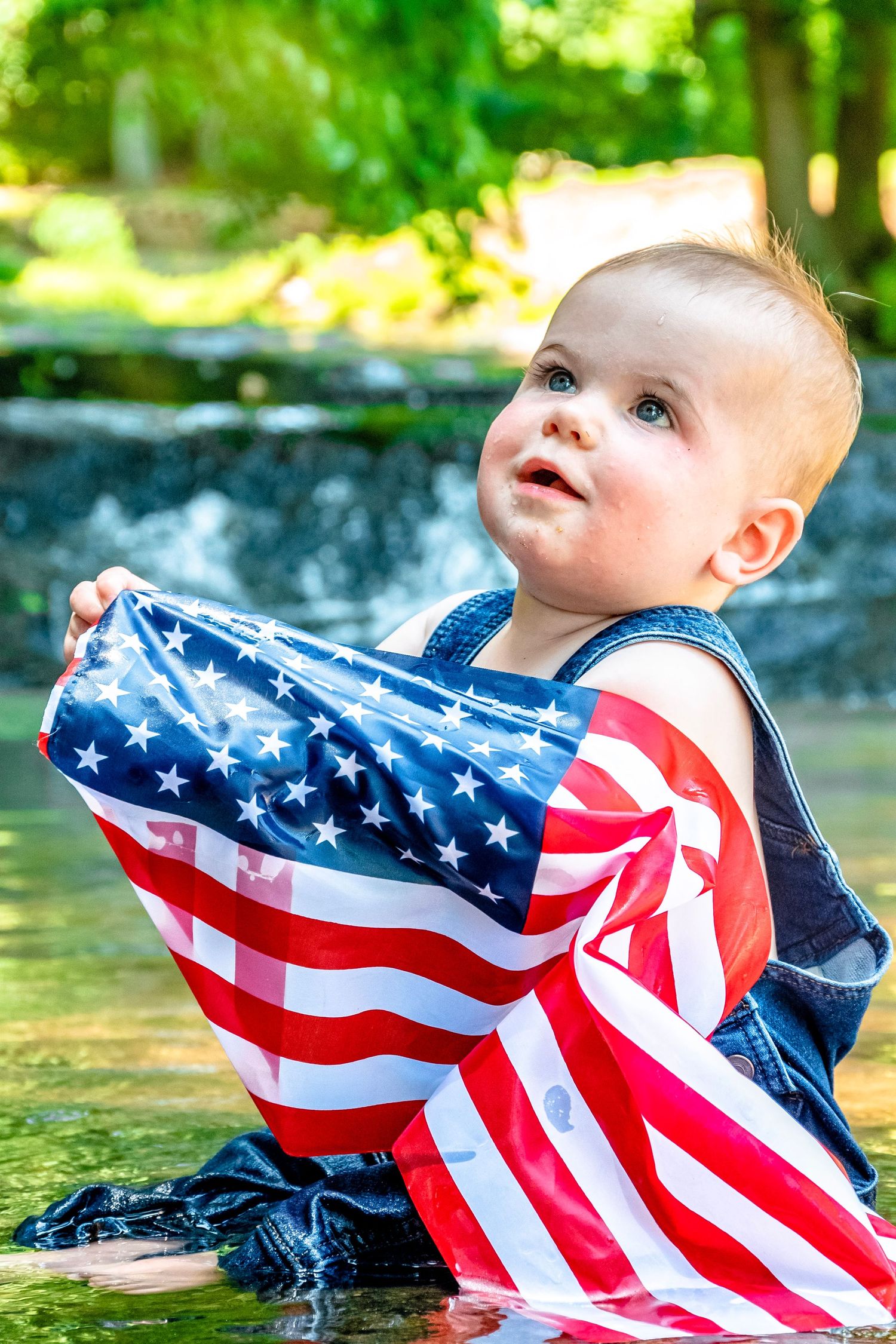 Adorable baby in denim overalls holding an American flag while sitting in water outdoors.
