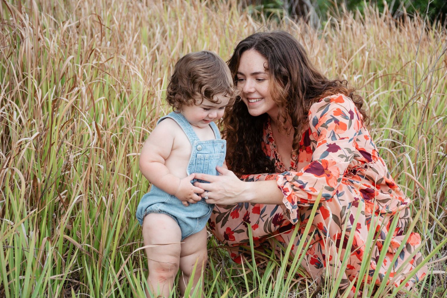 A smiling mother in a floral dress crouches with her toddler in blue overalls among tall golden grasses.