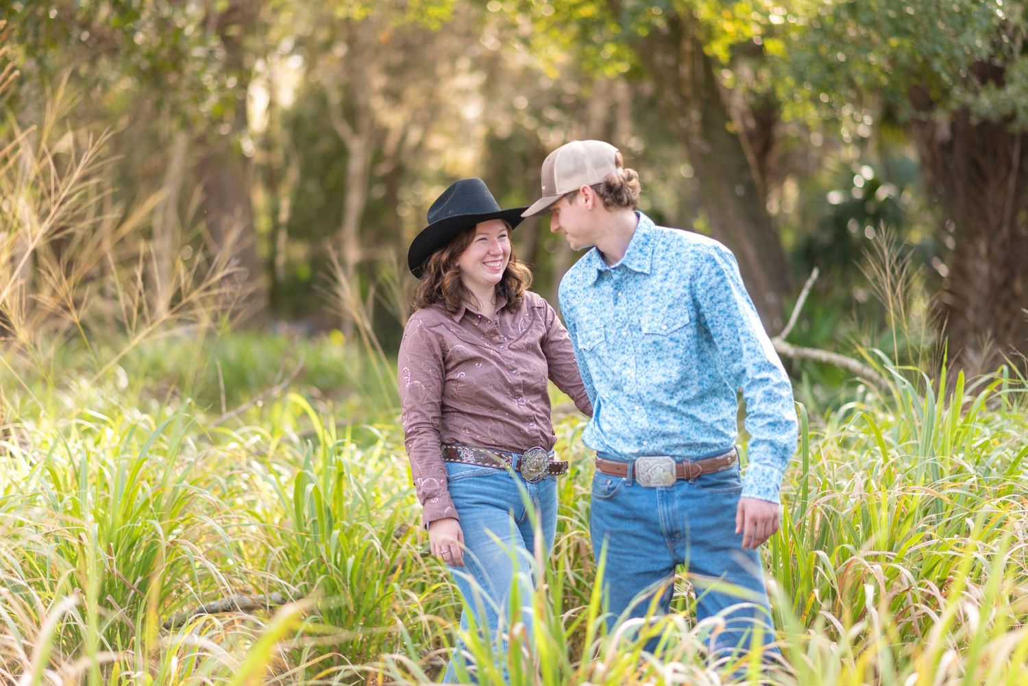 Couple in cowboy hats and jeans smiling at each other among tall green grass in a lush outdoor setting.