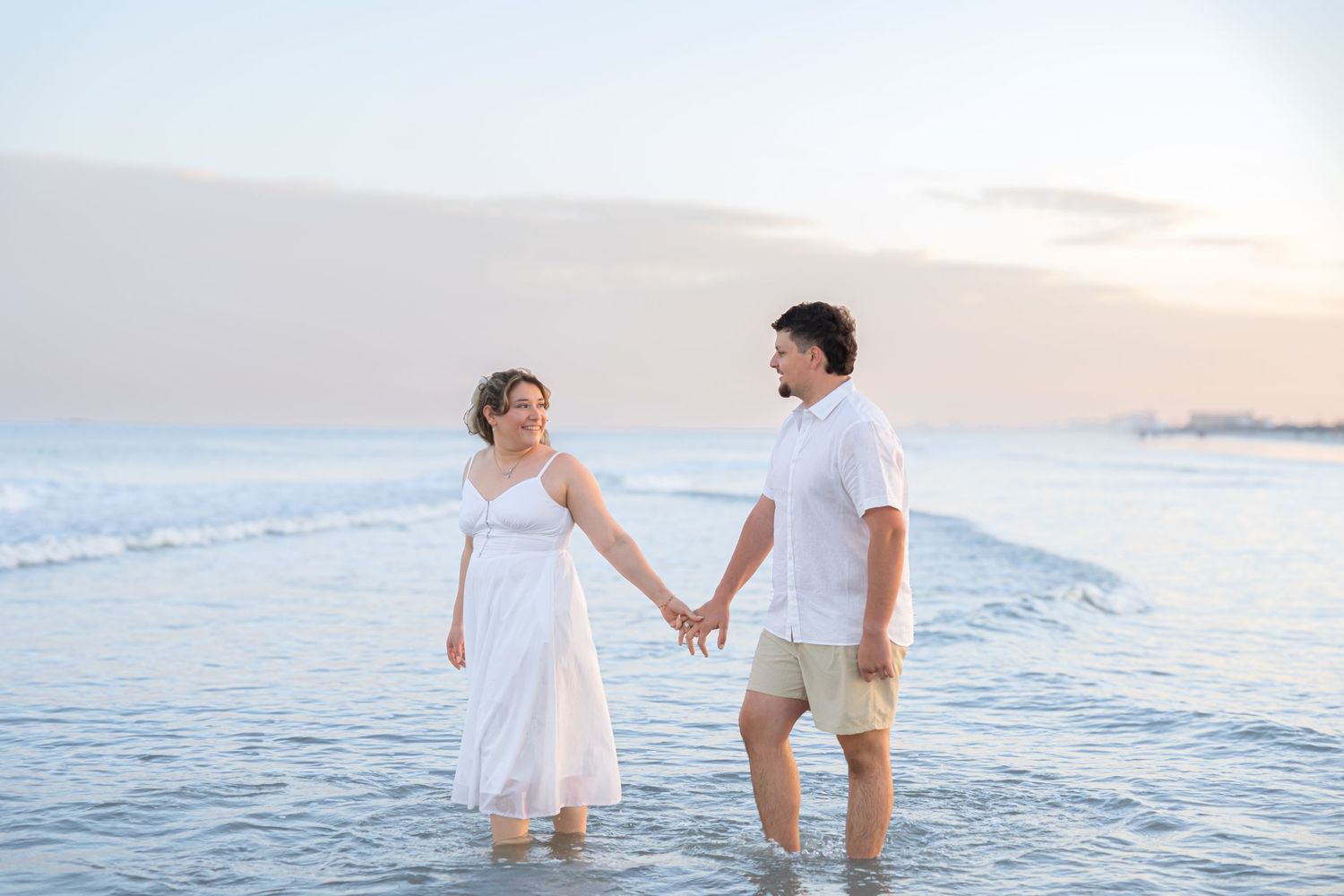 Couple holding hands wading in ocean waves at sunset, woman in white dress, man in white shirt and khaki shorts.