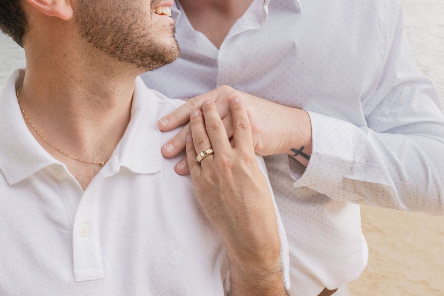 Two men in white shirts share a tender moment, one resting a ringed hand on the other's shoulder.