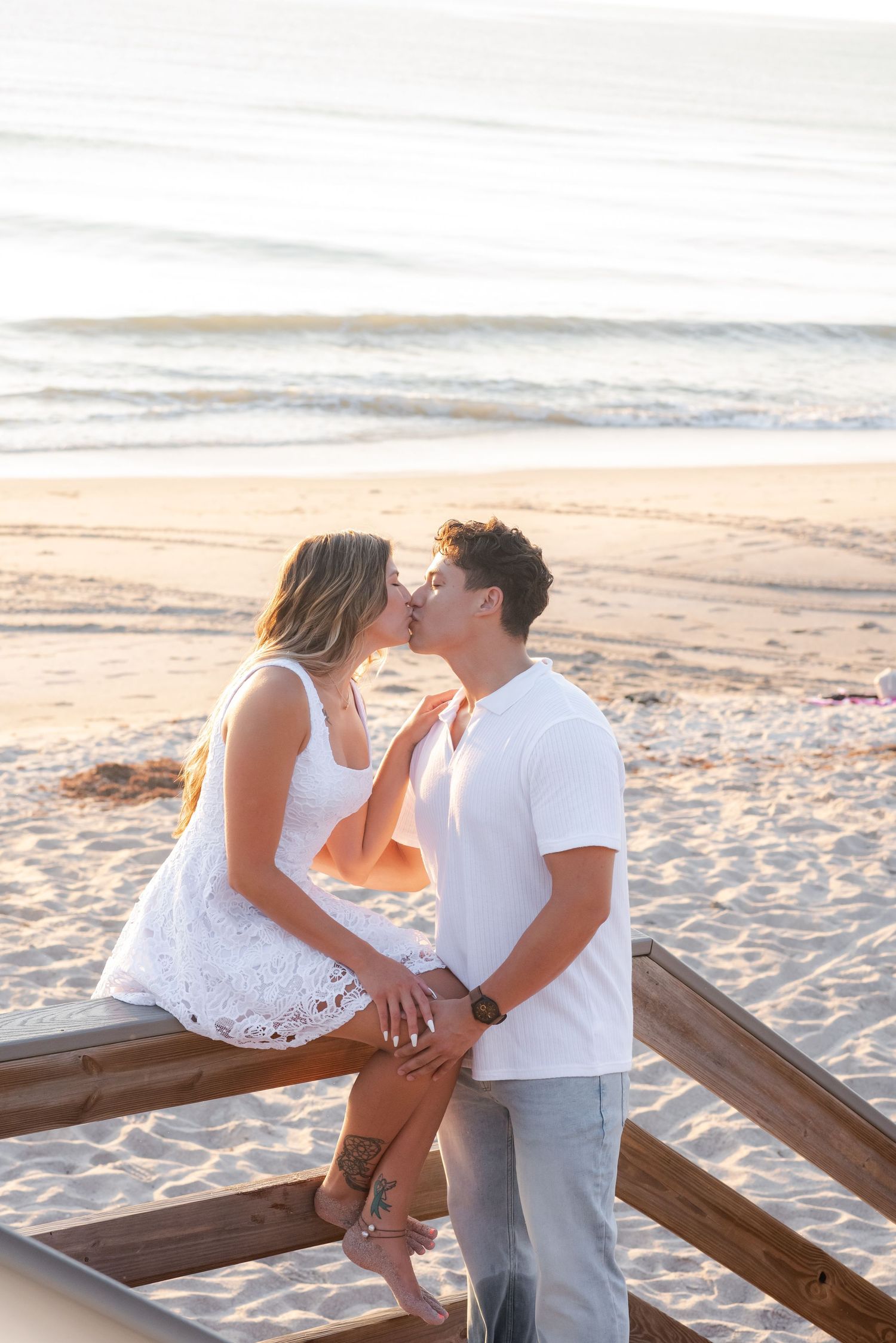 Couple sharing a romantic kiss on a wooden beach railing at sunset, wearing white outfits with ocean waves behind them.