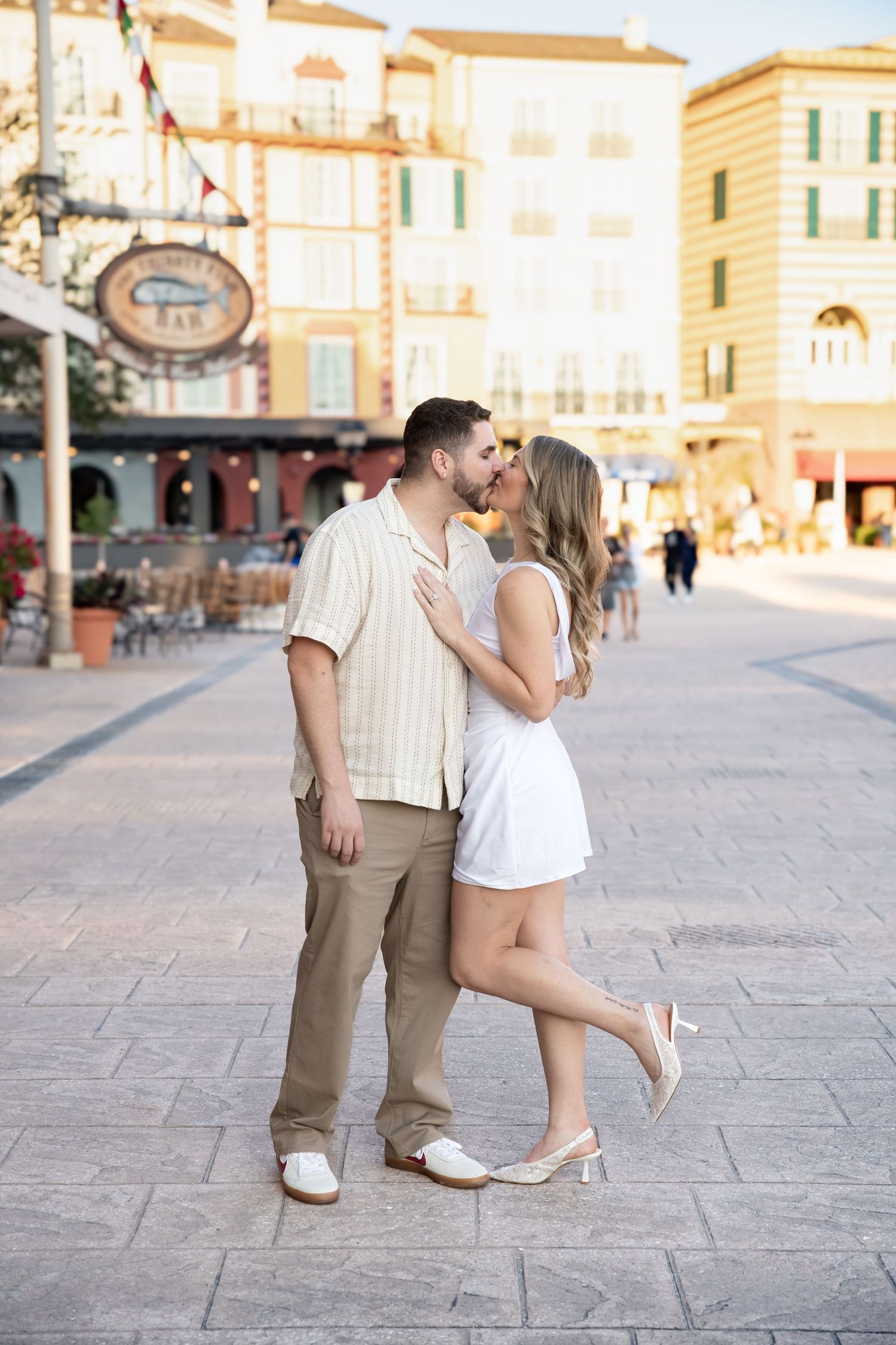 Couple kissing in a sunlit European-style plaza with colorful buildings and outdoor cafe.