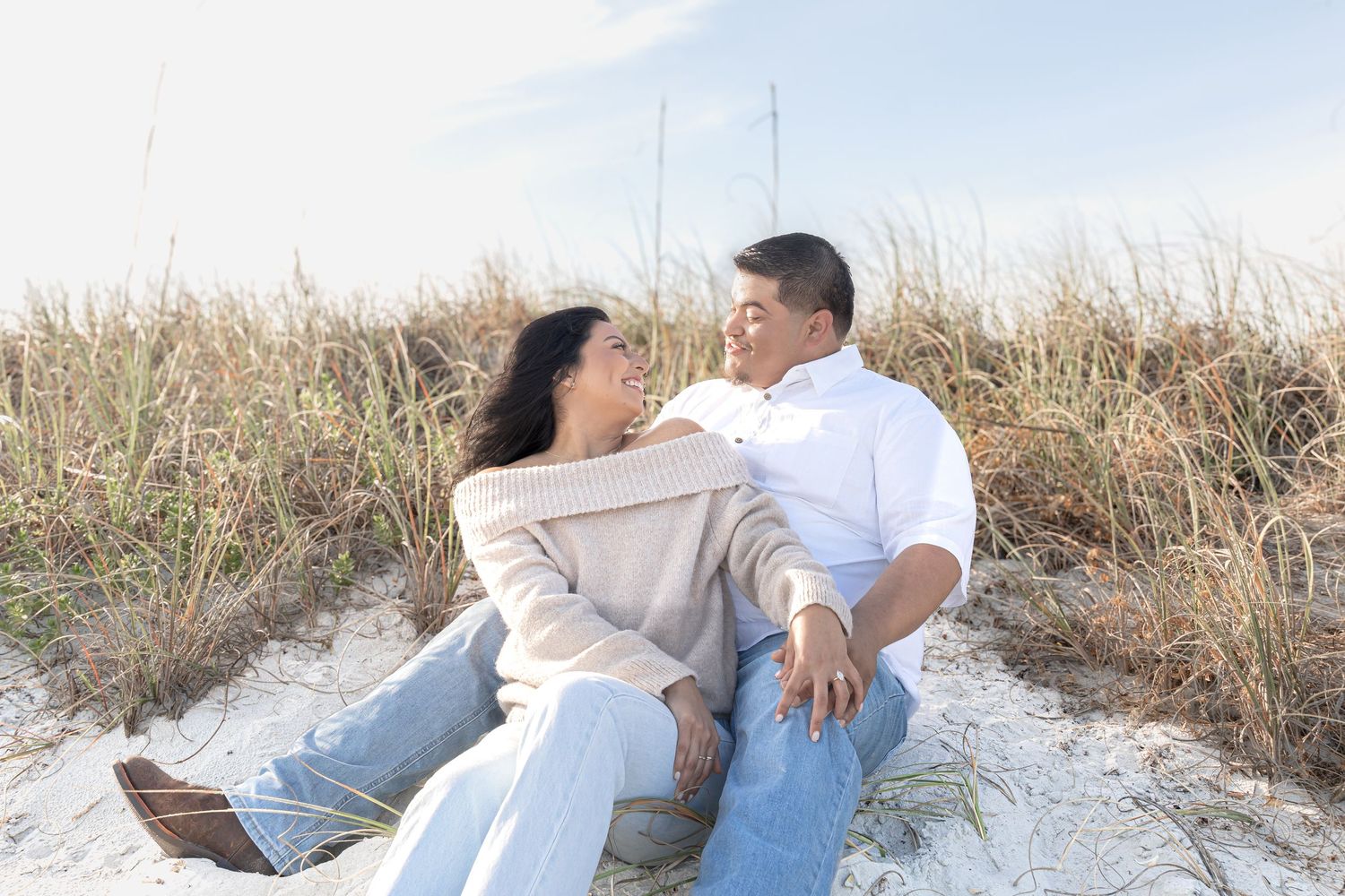 Couple sitting together on sandy beach dunes, smiling at each other, surrounded by tall grass on a sunny day.