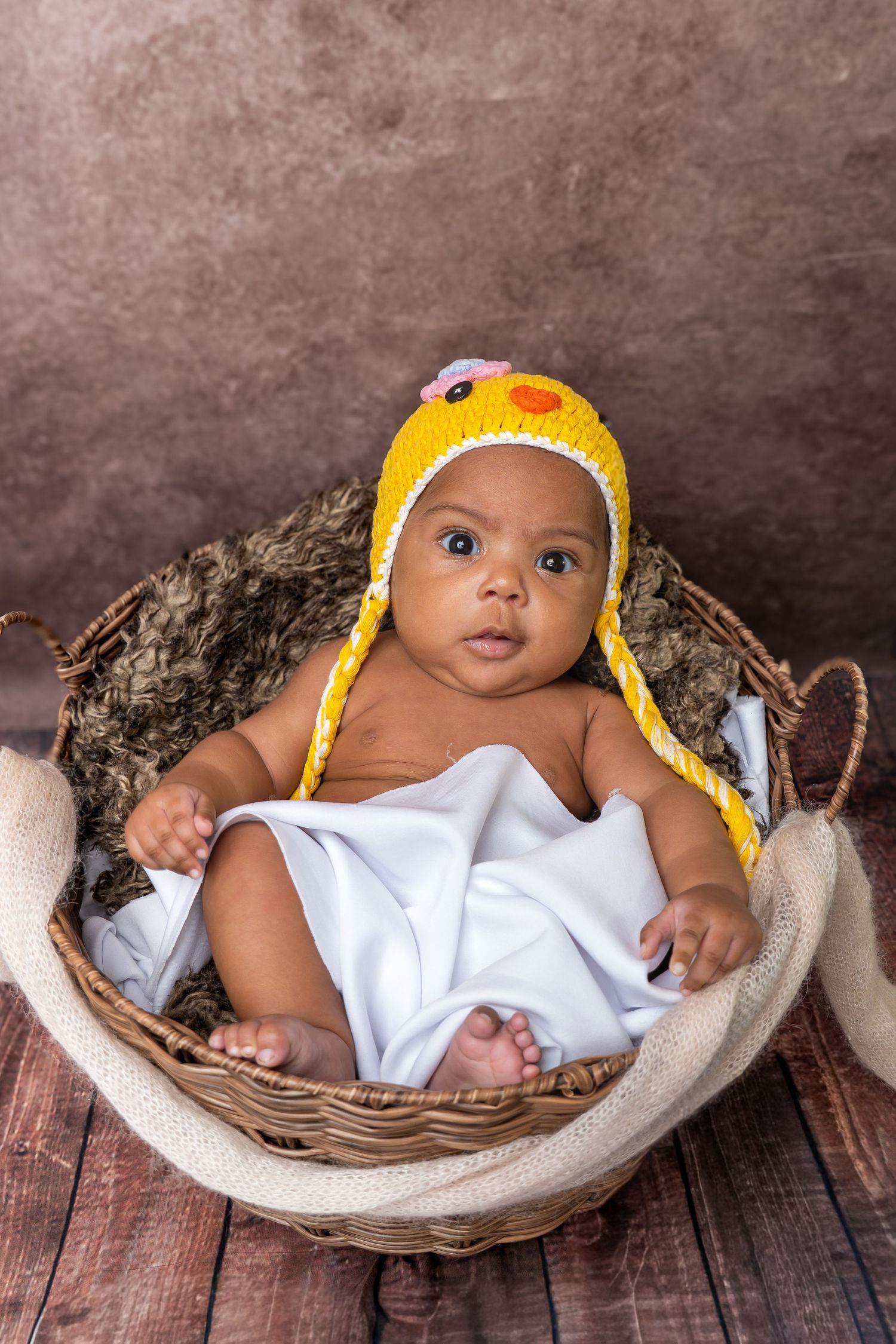Cute infant in yellow knit chick hat sitting upright in wicker basket with white blanket on wooden floor.