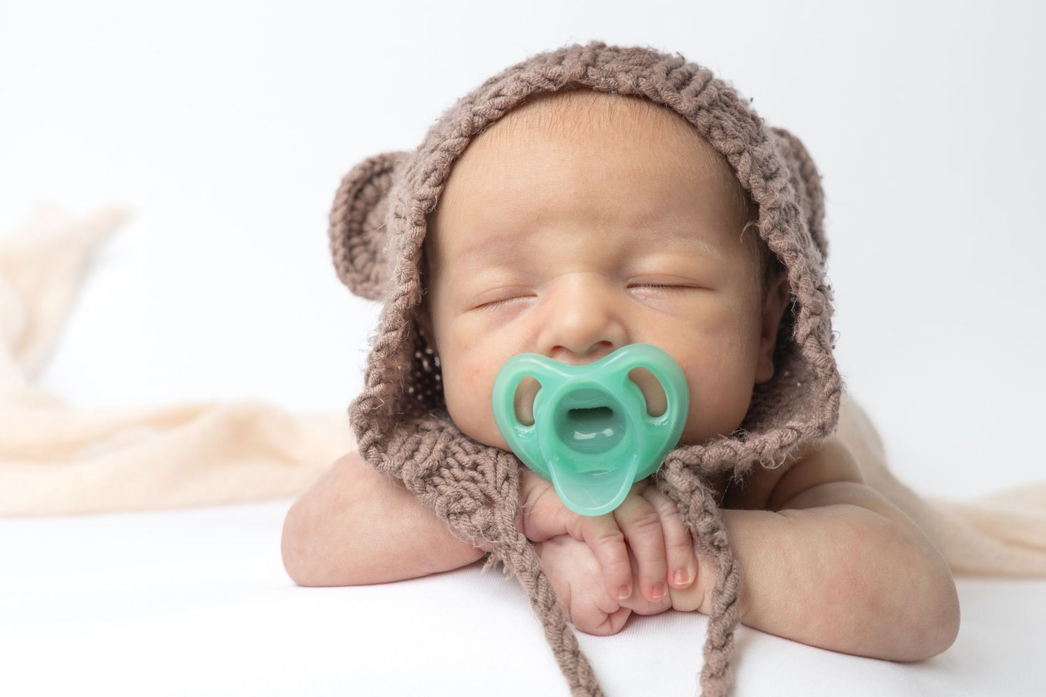 Sleeping newborn baby wearing a brown knitted bear hat with a teal pacifier, posed on a white background.