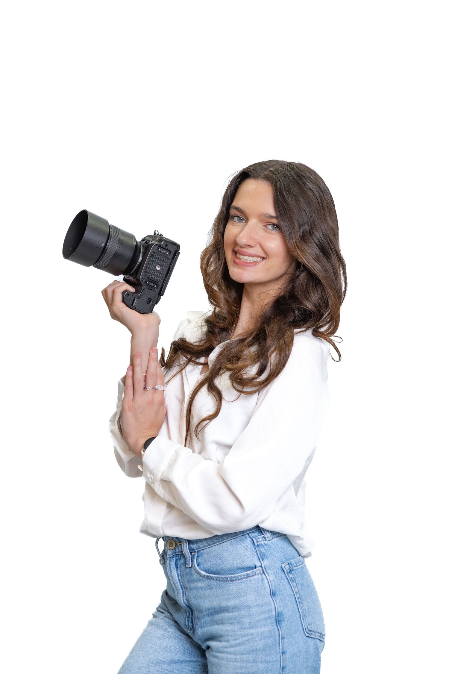 Smiling female photographer holding a professional DSLR camera, wearing white blouse and jeans on white background.
