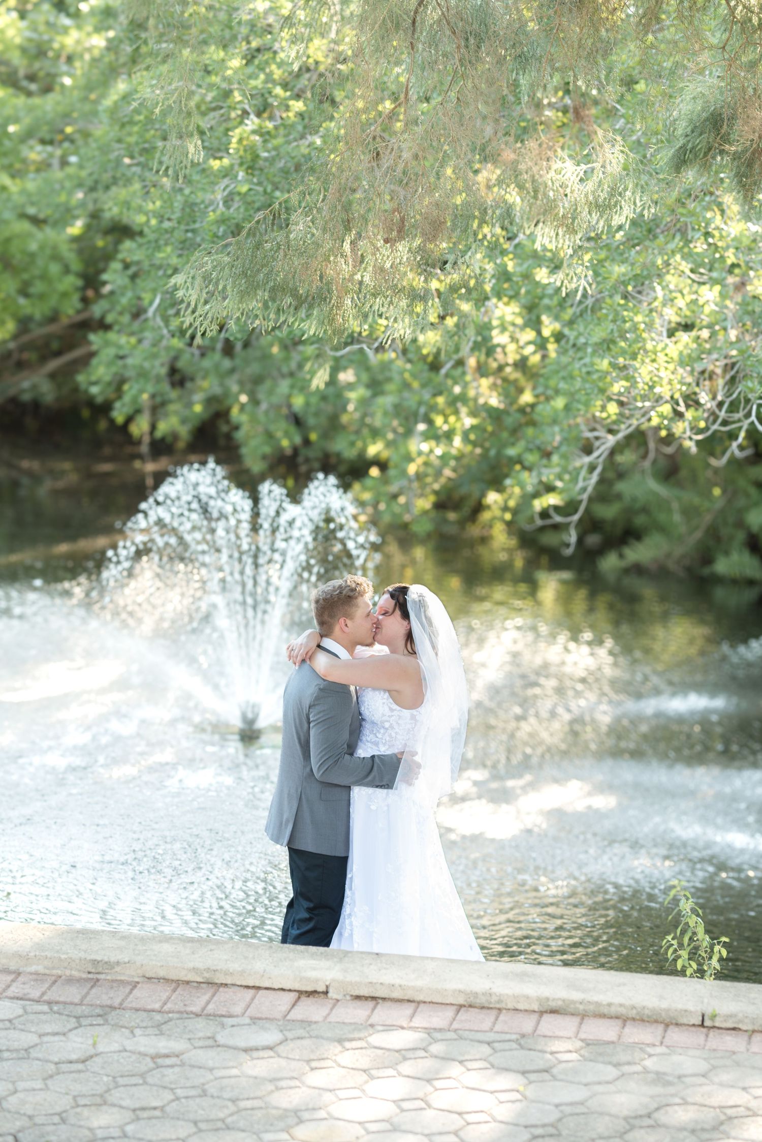 Newlyweds sharing a romantic kiss near a garden fountain surrounded by lush green trees.