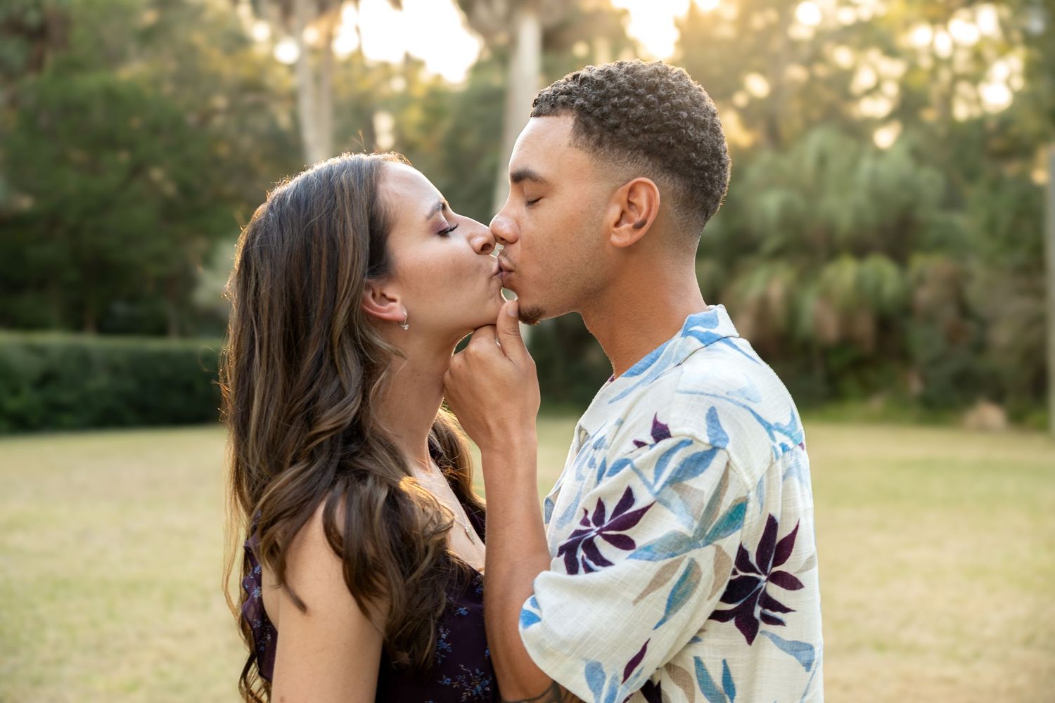 A couple shares a romantic kiss outdoors in a lush green park setting during golden hour.