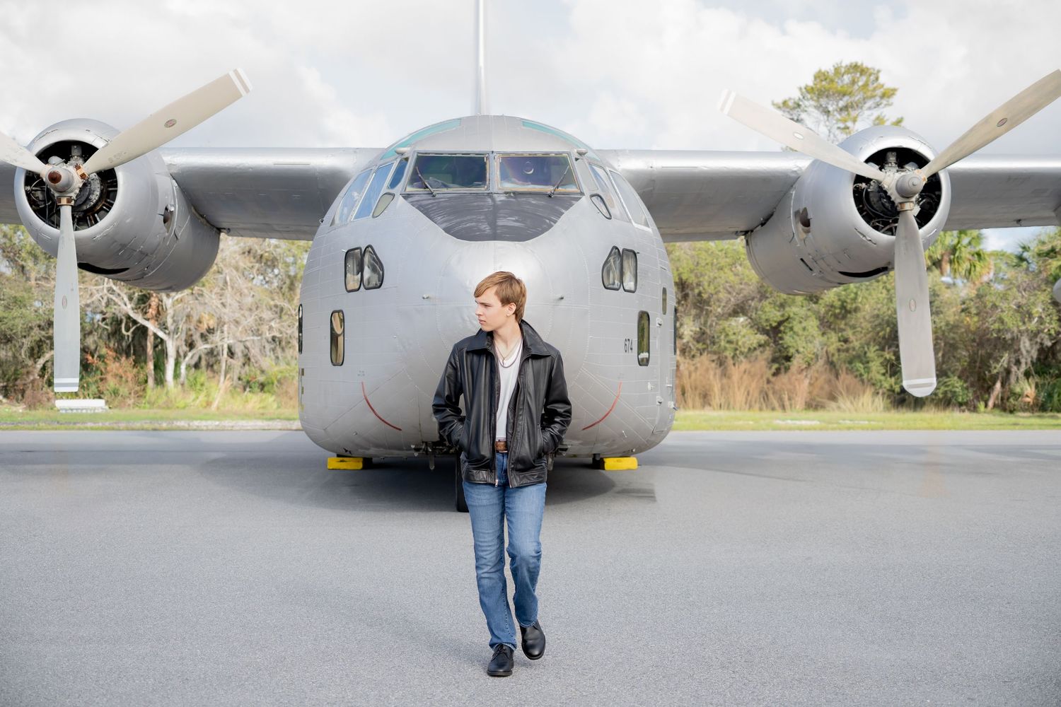Young woman in leather jacket and jeans standing confidently in front of a large military cargo aircraft on a runway.