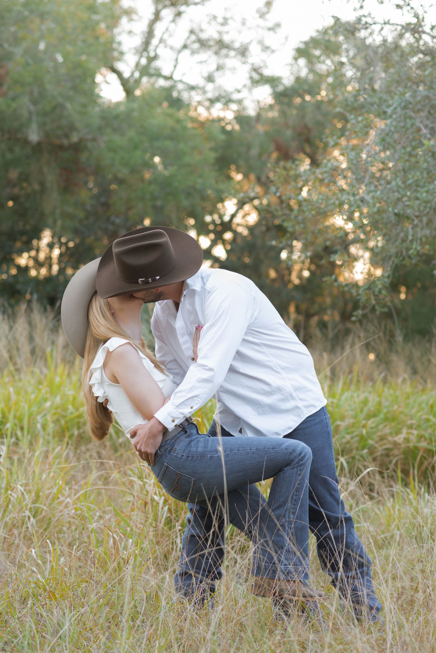 Couple sharing a romantic dip kiss in a golden meadow, wearing cowboy hats, jeans, and boots with trees in background.